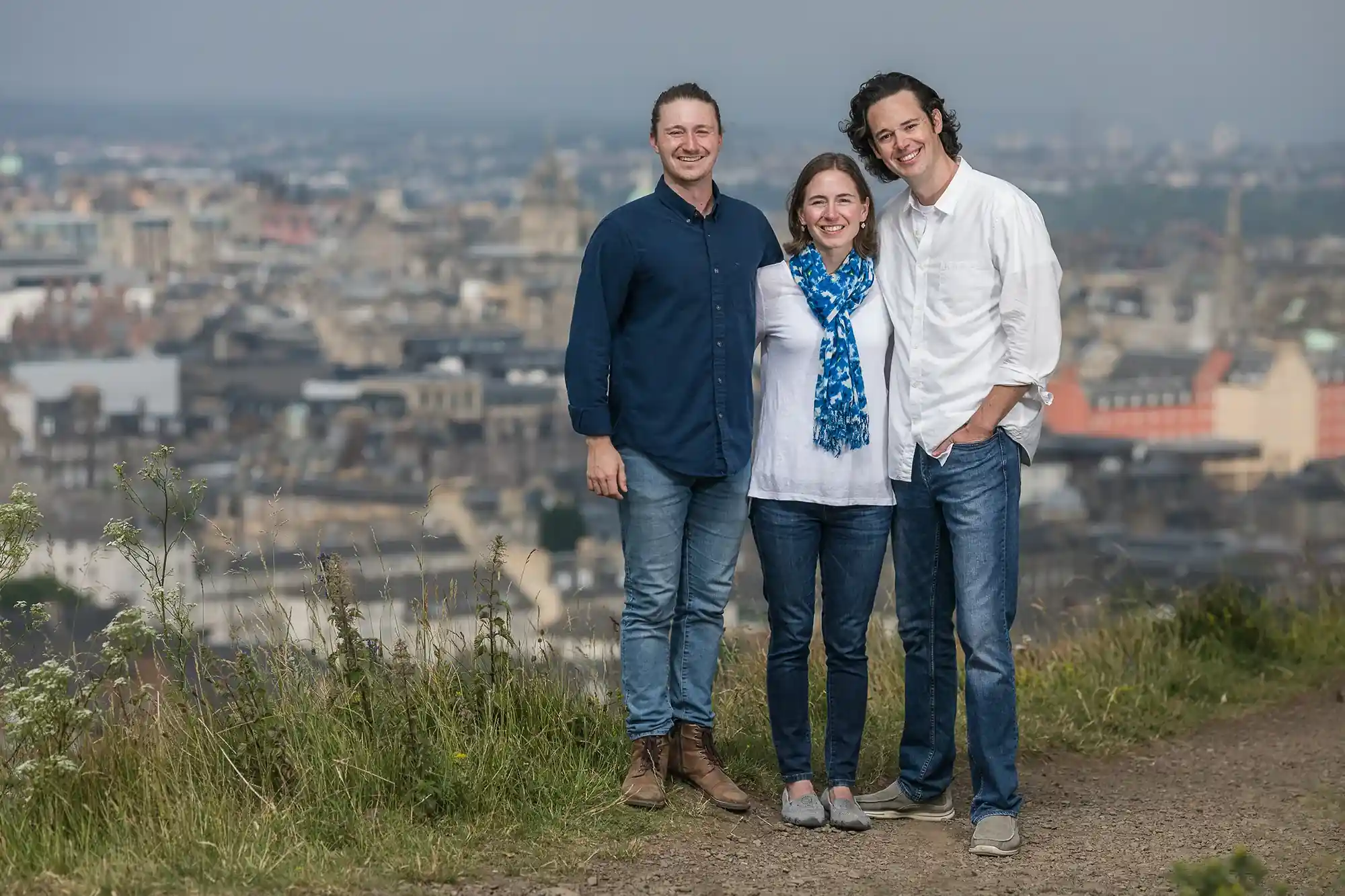 Three people stand smiling on a hilltop with a cityscape in the background. Two men and a woman, all casually dressed, pose together on a slightly overcast day.