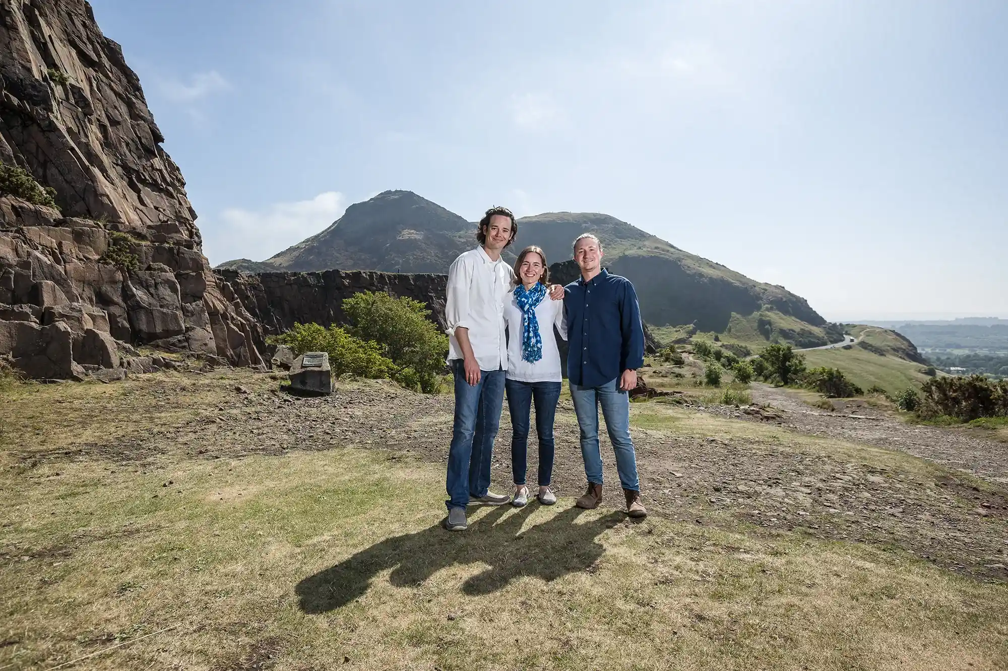 Three people stand closely together smiling, with rocky hills and a clear sky in the background.