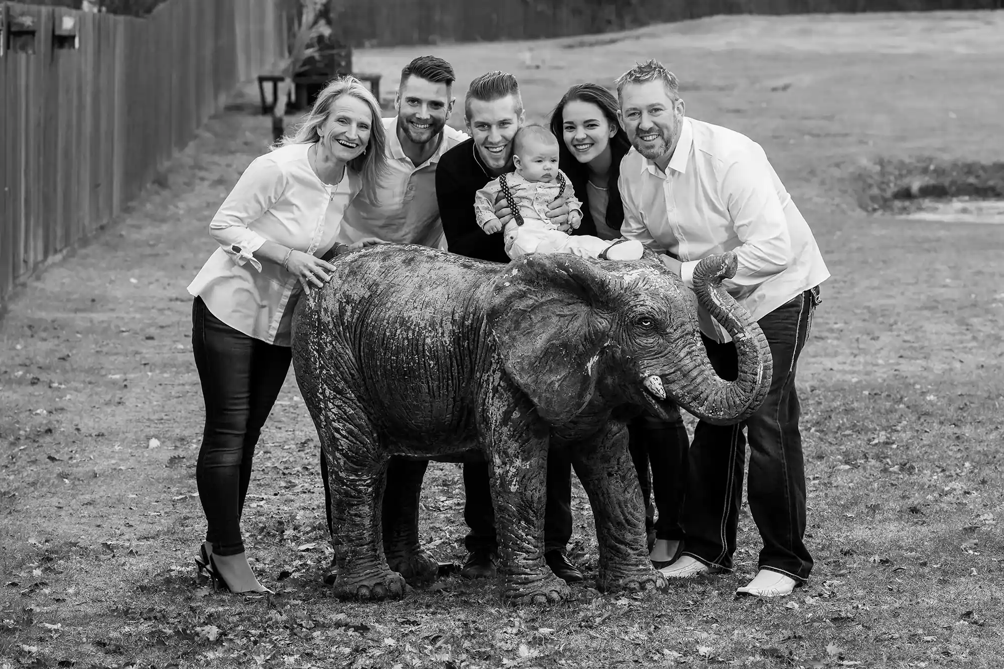 Black and white photo of five adults and a baby posing with a baby elephant outdoors. Everyone is smiling and standing close together.