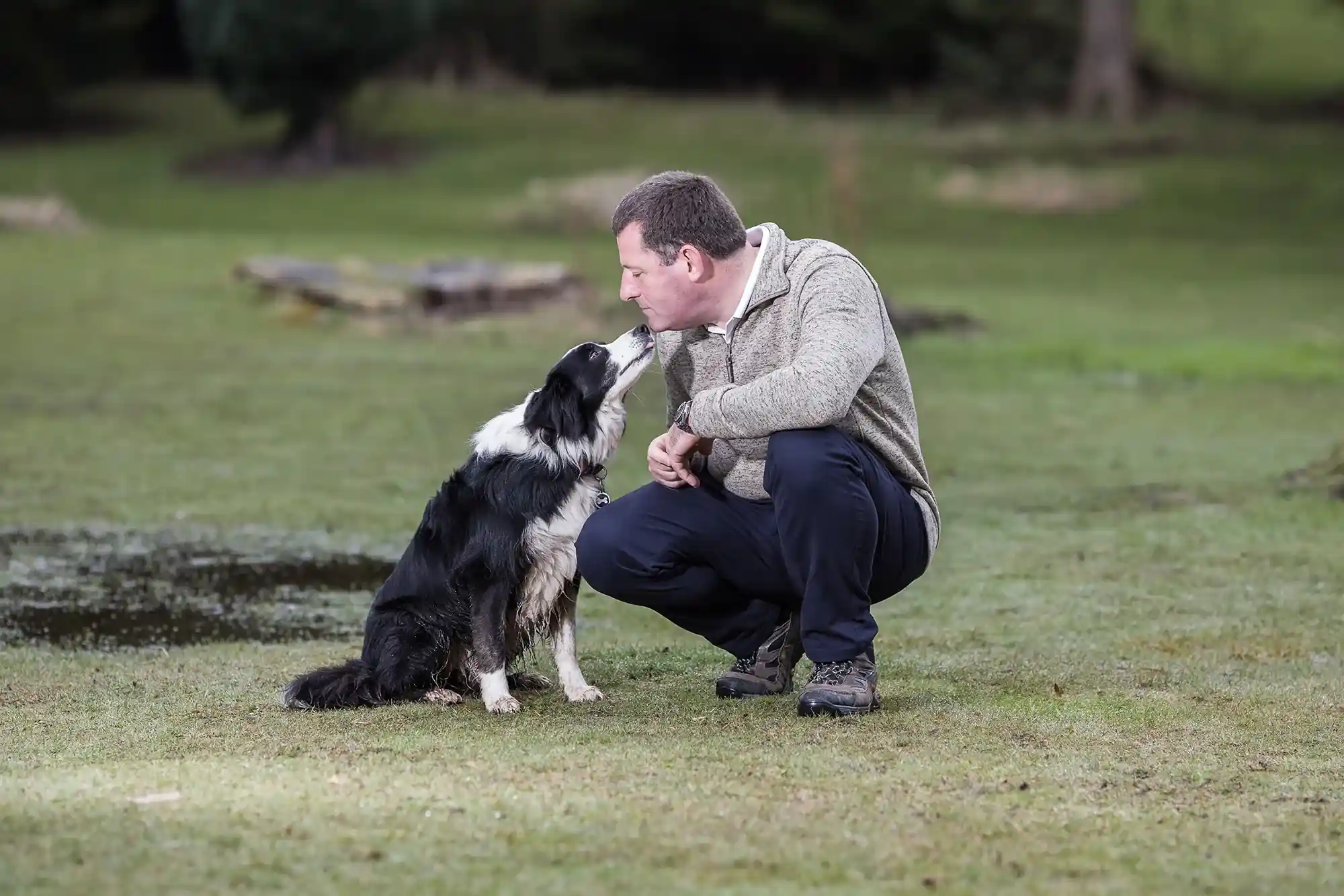 A man in a sweater and dark pants squats on a grassy field, touching noses with a black and white dog.