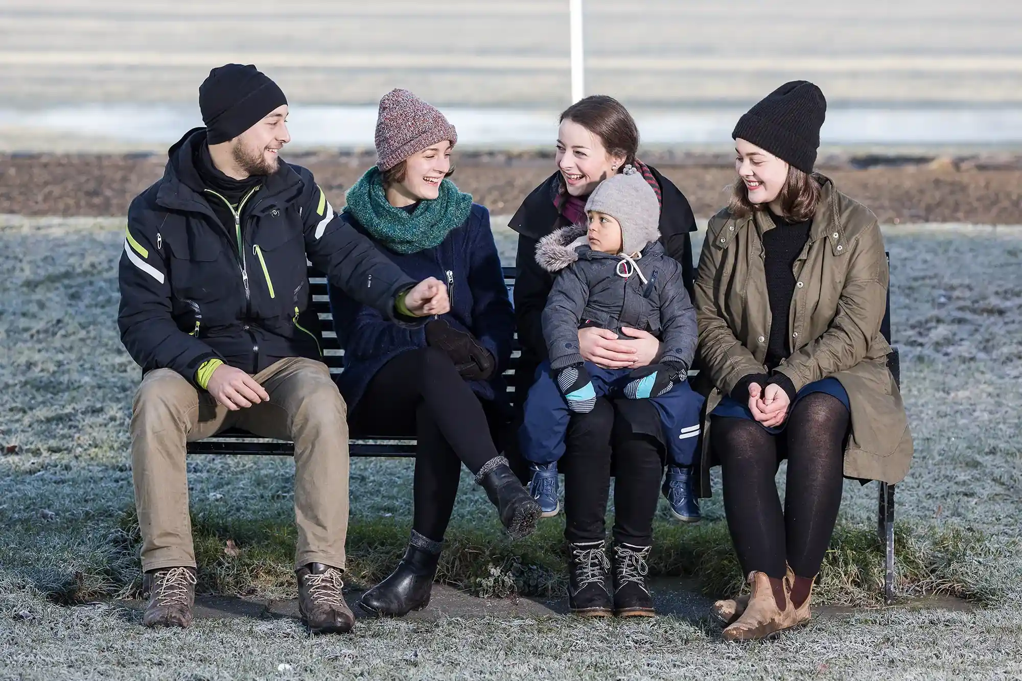 Five people, including one young child, sitting on a bench outdoors during winter. All are dressed in winter clothing, smiling and engaged in conversation.