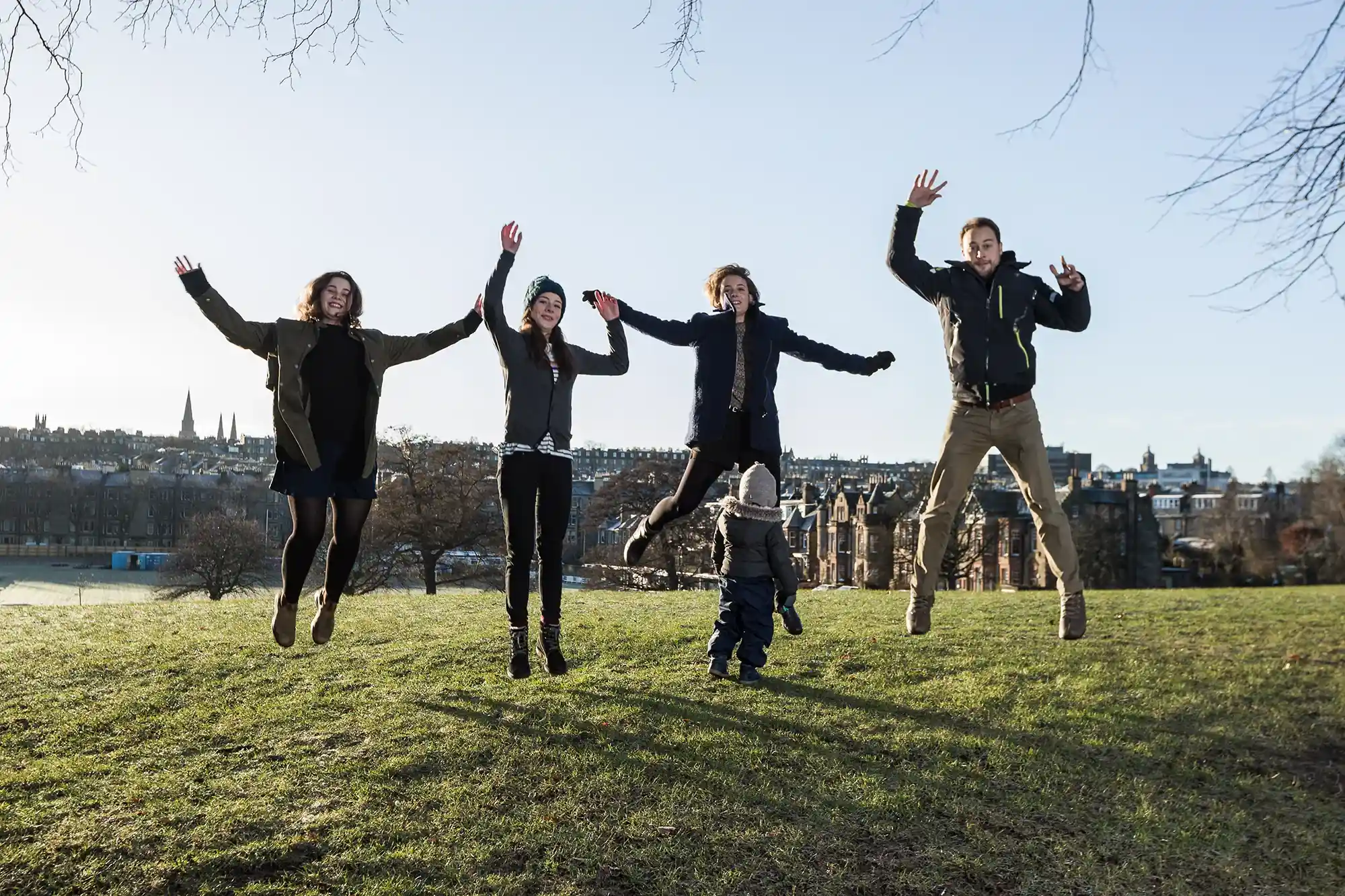 Four adults and one child are mid-jump in a park on a clear day, with a cityscape in the background.