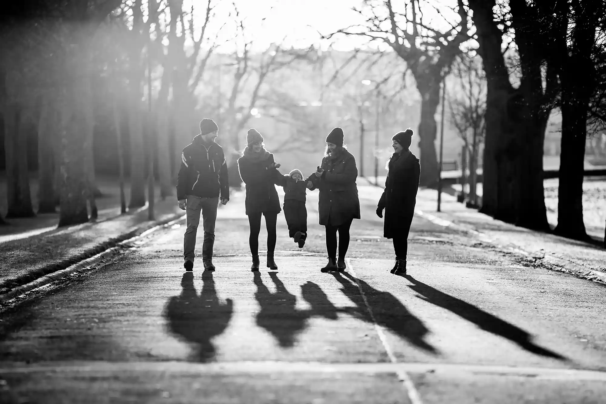 A group of five people walking down a tree-lined path in winter, casting long shadows in the late afternoon sun. The scene is in black and white.