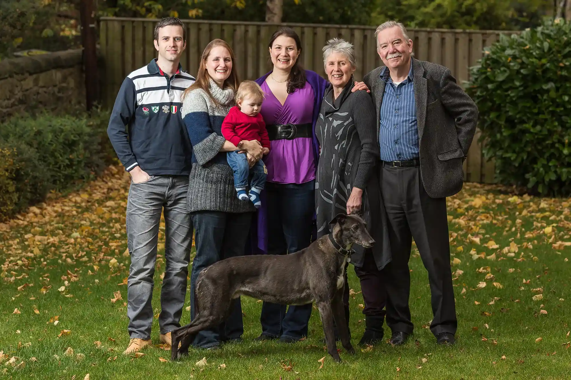 A group of six people, including a child, and a dog pose for a photo on a grassy lawn with a wooden fence and leafy trees in the background.