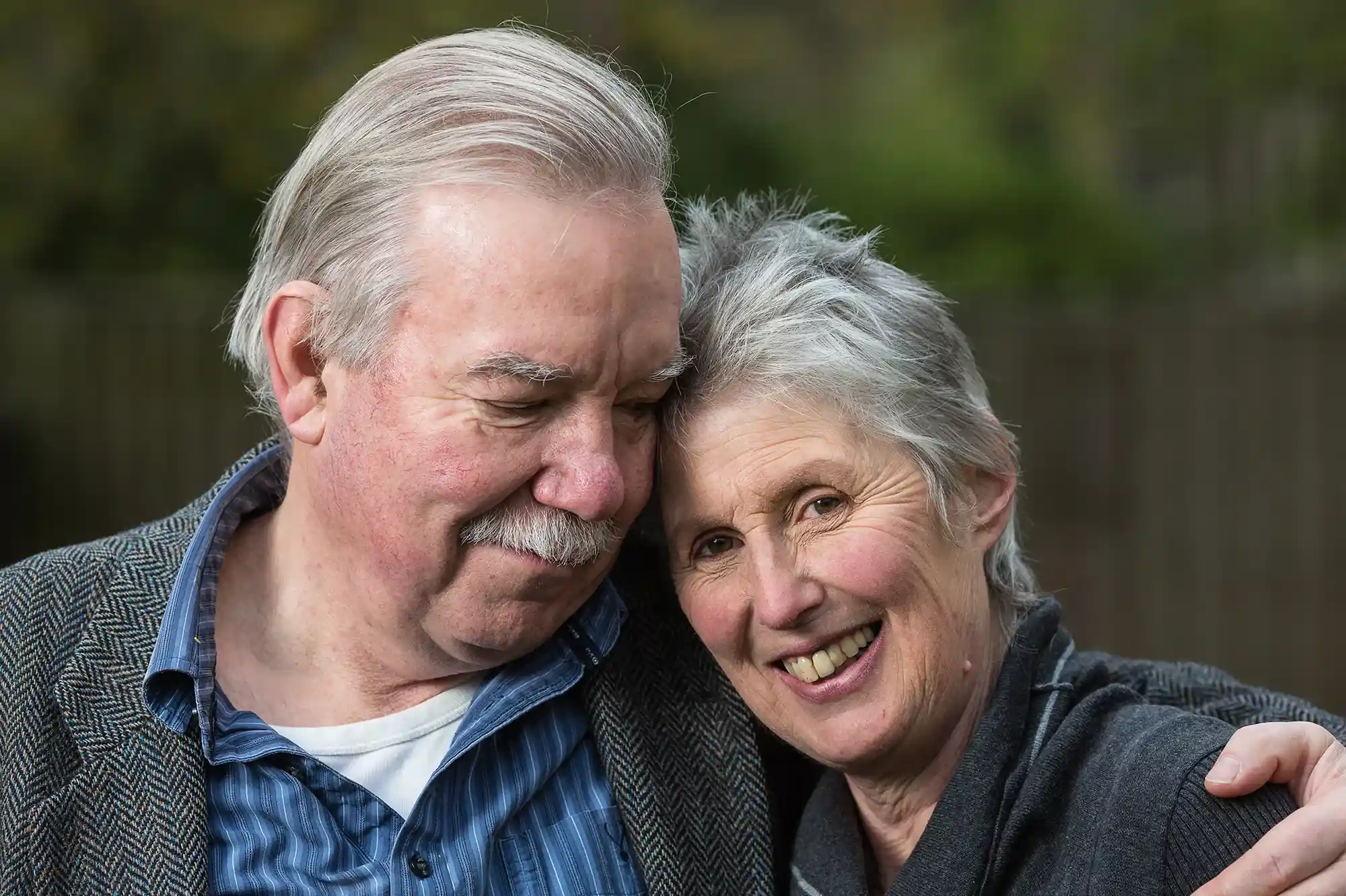 An elderly couple stands close together outdoors, smiling warmly. The woman has short gray hair and the man has white hair and a mustache. They are dressed in casual, warm clothing.