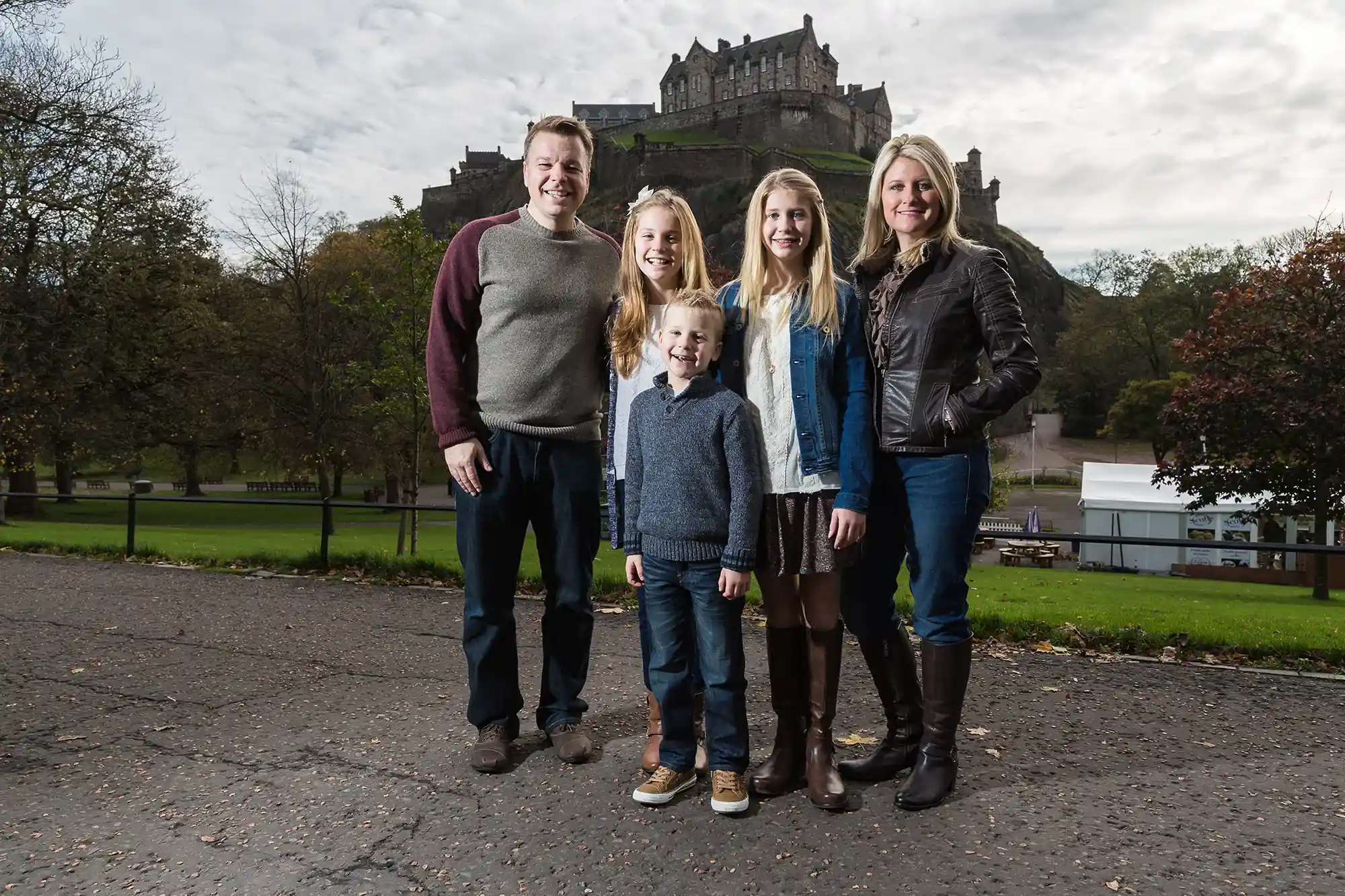 A family of five stands on a paved path with greenery and a large castle in the background. The group includes two adults and three children, all dressed in casual clothing.