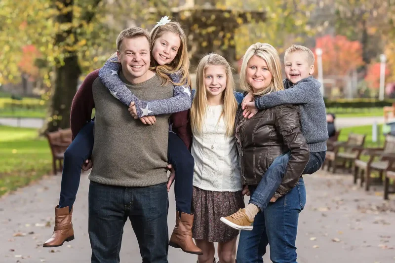 A group of five stands in a park with autumn foliage. Two children are carried on the adults' backs, while a third child stands in between them. They are all smiling at the camera.