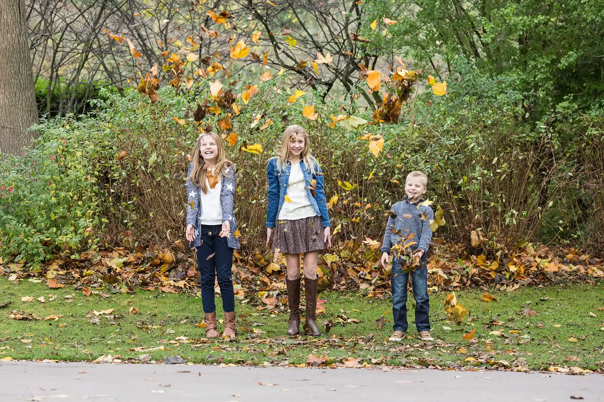 Three children stand on grass, smiling as they throw autumn leaves in the air, with bushes and trees in the background.
