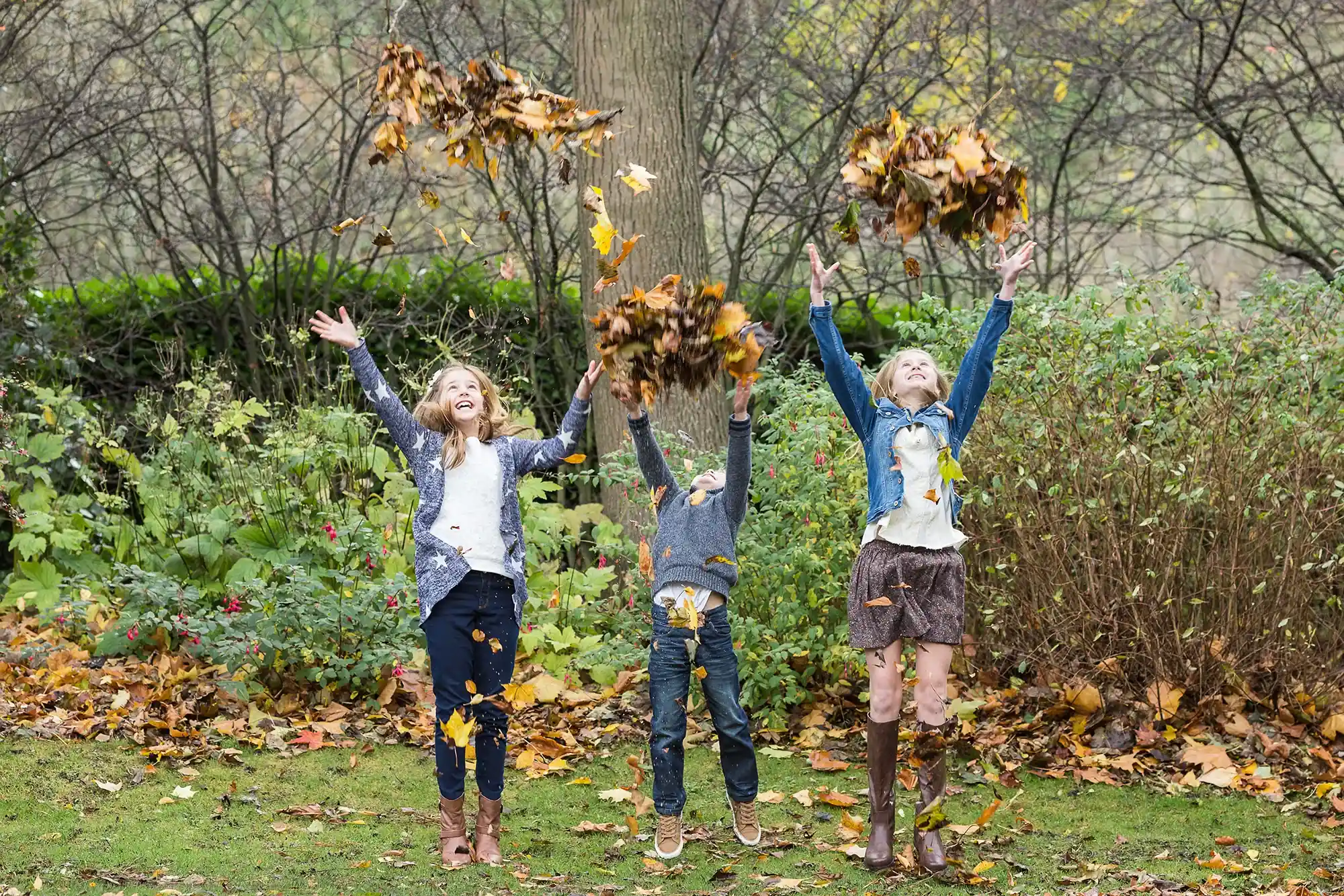 Three children are playing outdoors, tossing autumn leaves into the air. Trees and bushes form the background.