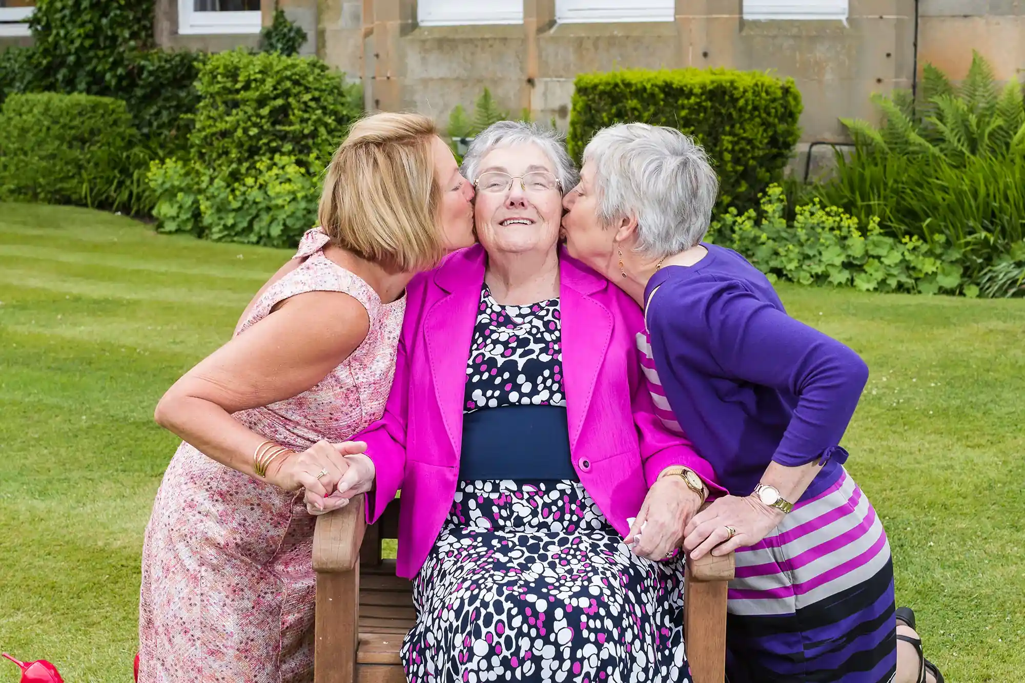 Three elderly women are sitting on a wooden bench in a garden. The woman in the center is being kissed on the cheeks by the women on either side. All are smiling.