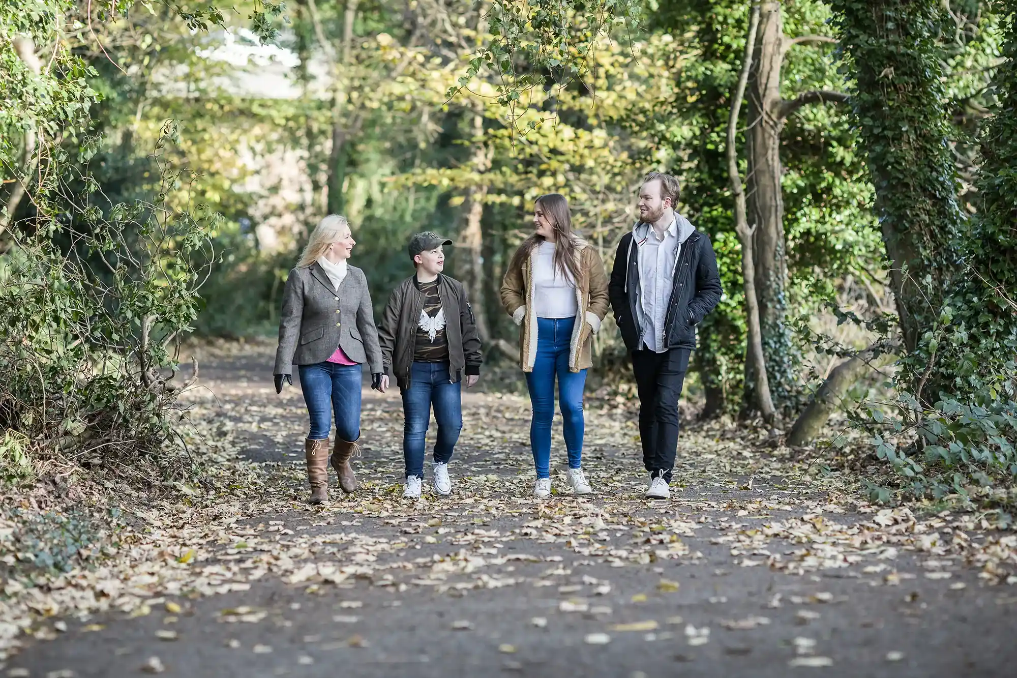 Four people walking together on a forest path surrounded by autumn leaves and trees, engaging in conversation and appearing relaxed.