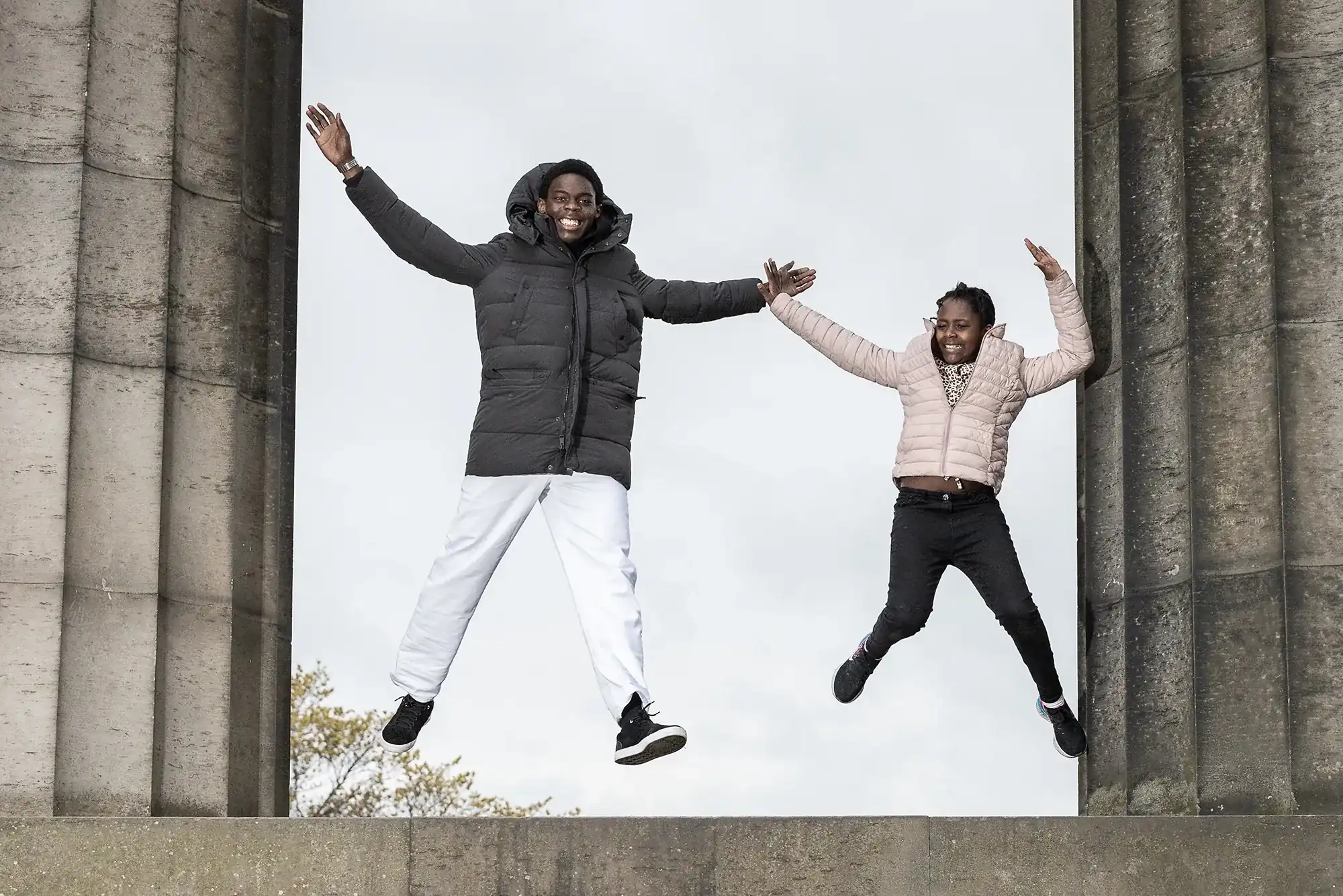 Two people are jumping and holding hands in front of a stone structure. They are both wearing jackets and appear to be smiling. The sky is overcast.