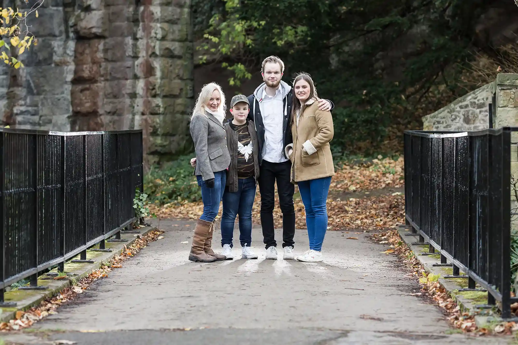 A group of four people, two adults and two children, stand together on a narrow path between railings, with greenery and a stone structure in the background.