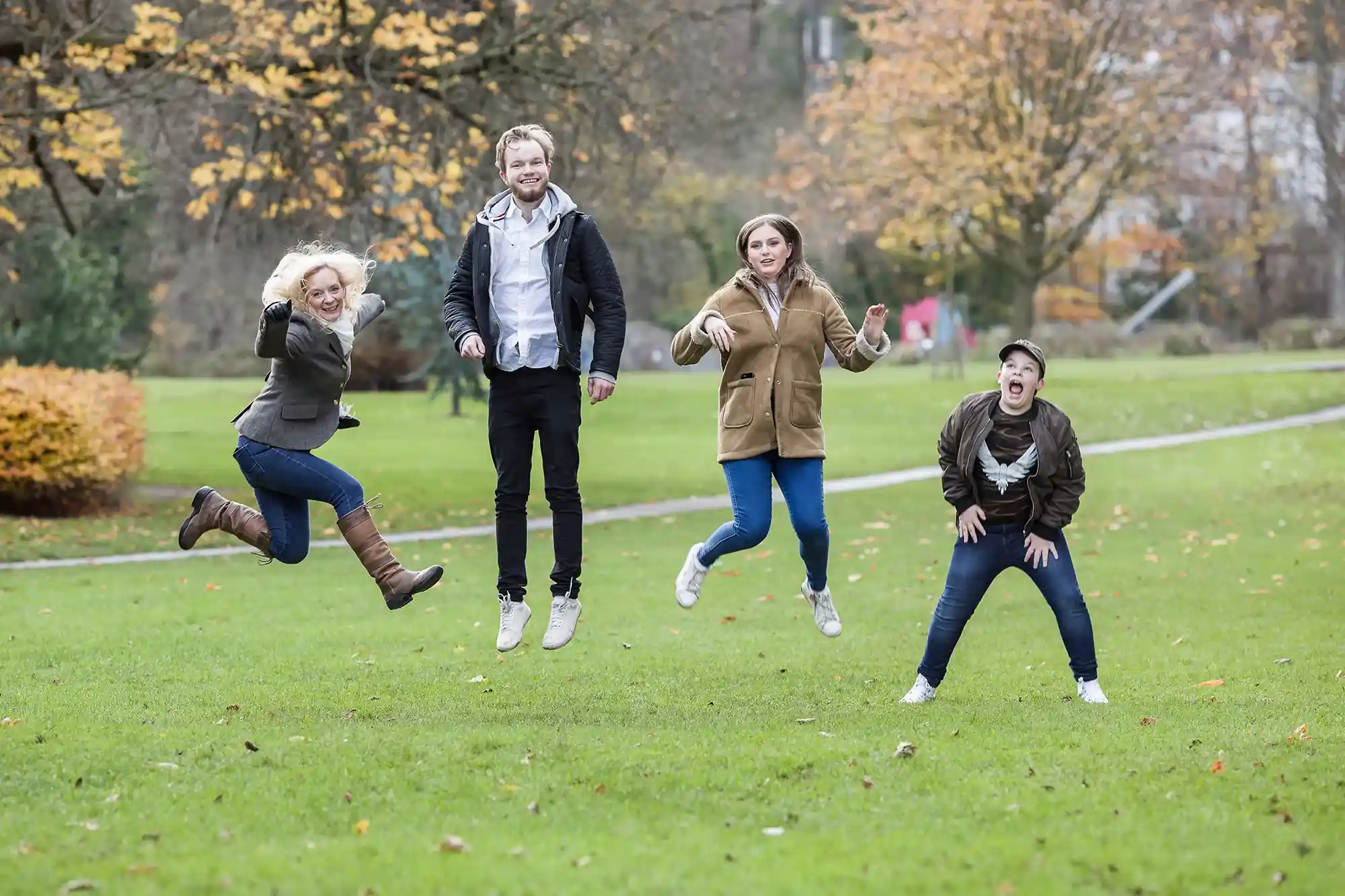 Four people jump in mid-air in a grassy park with autumn trees in the background. They are dressed in casual clothes and look cheerful.