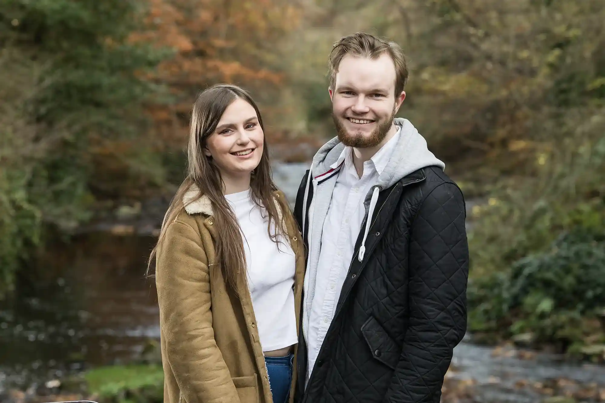 A man and woman stand outdoors near a flowing creek with autumn foliage in the background. Both are smiling at the camera.