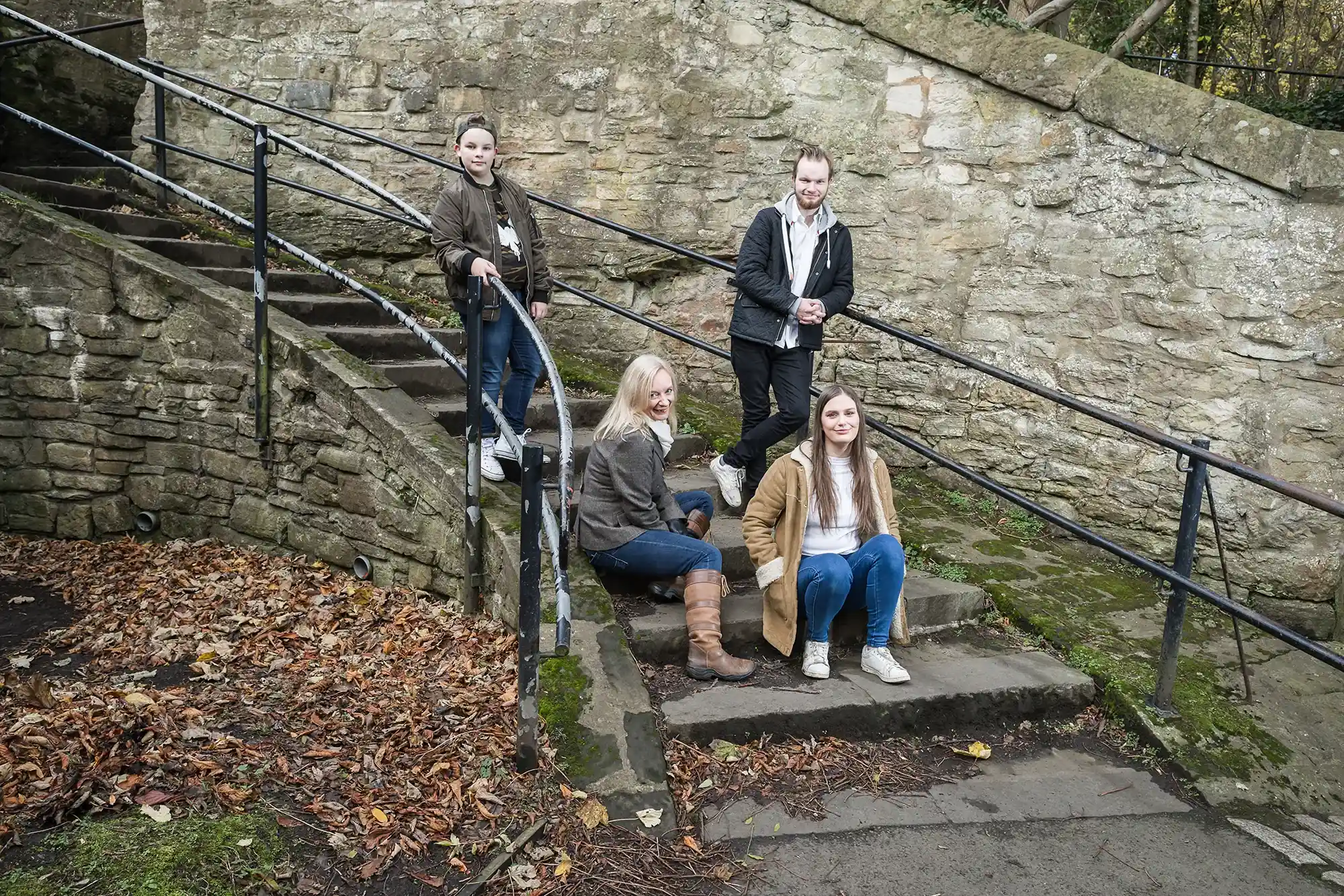 Four people pose on stone steps beside an old stone wall. Two women sit while two men stand, all dressed casually. Fallen autumn leaves cover parts of the ground.