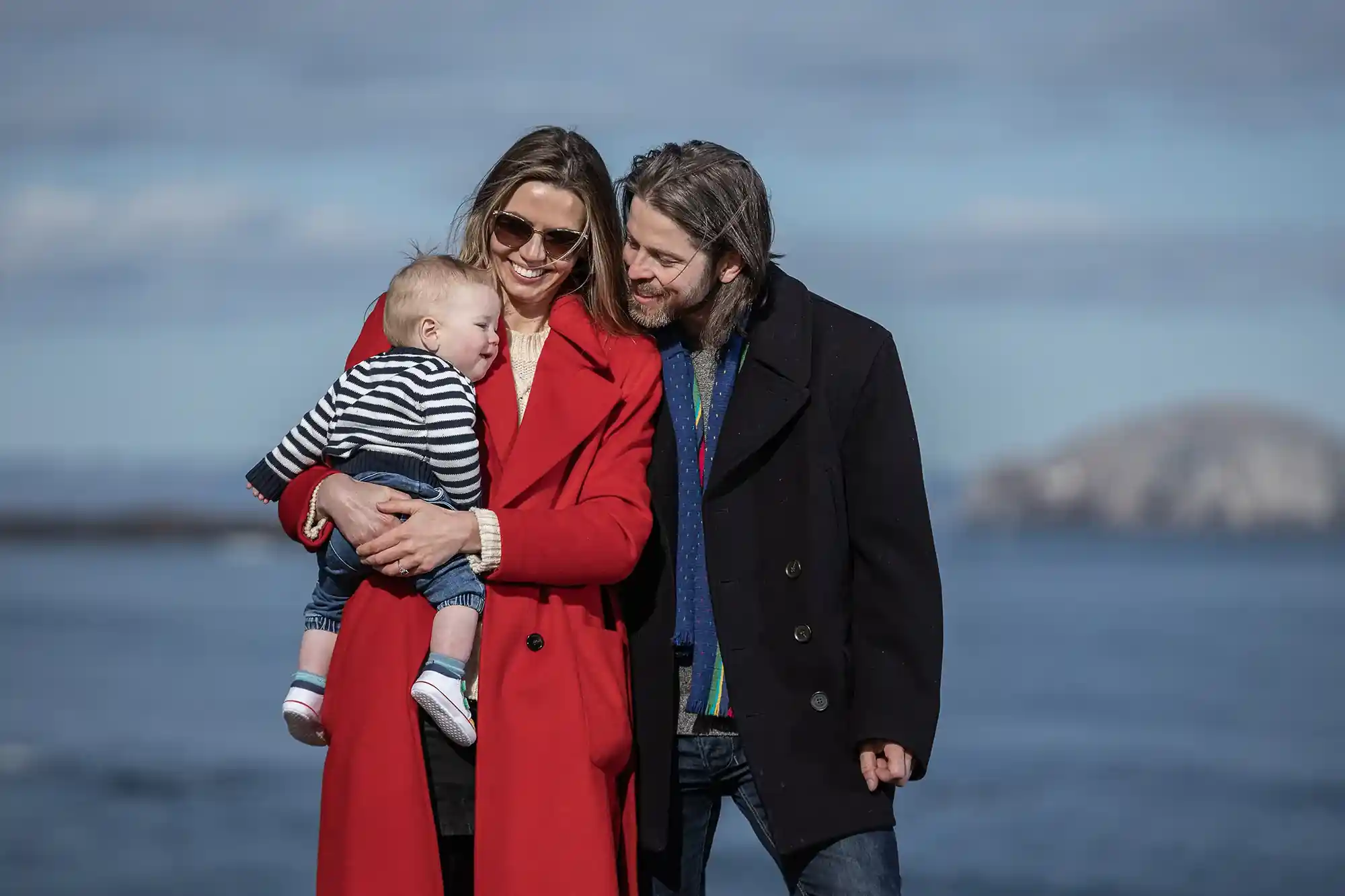 A woman in a red coat holds a baby next to a man in a black coat, all standing by a body of water with distant hills in the background.