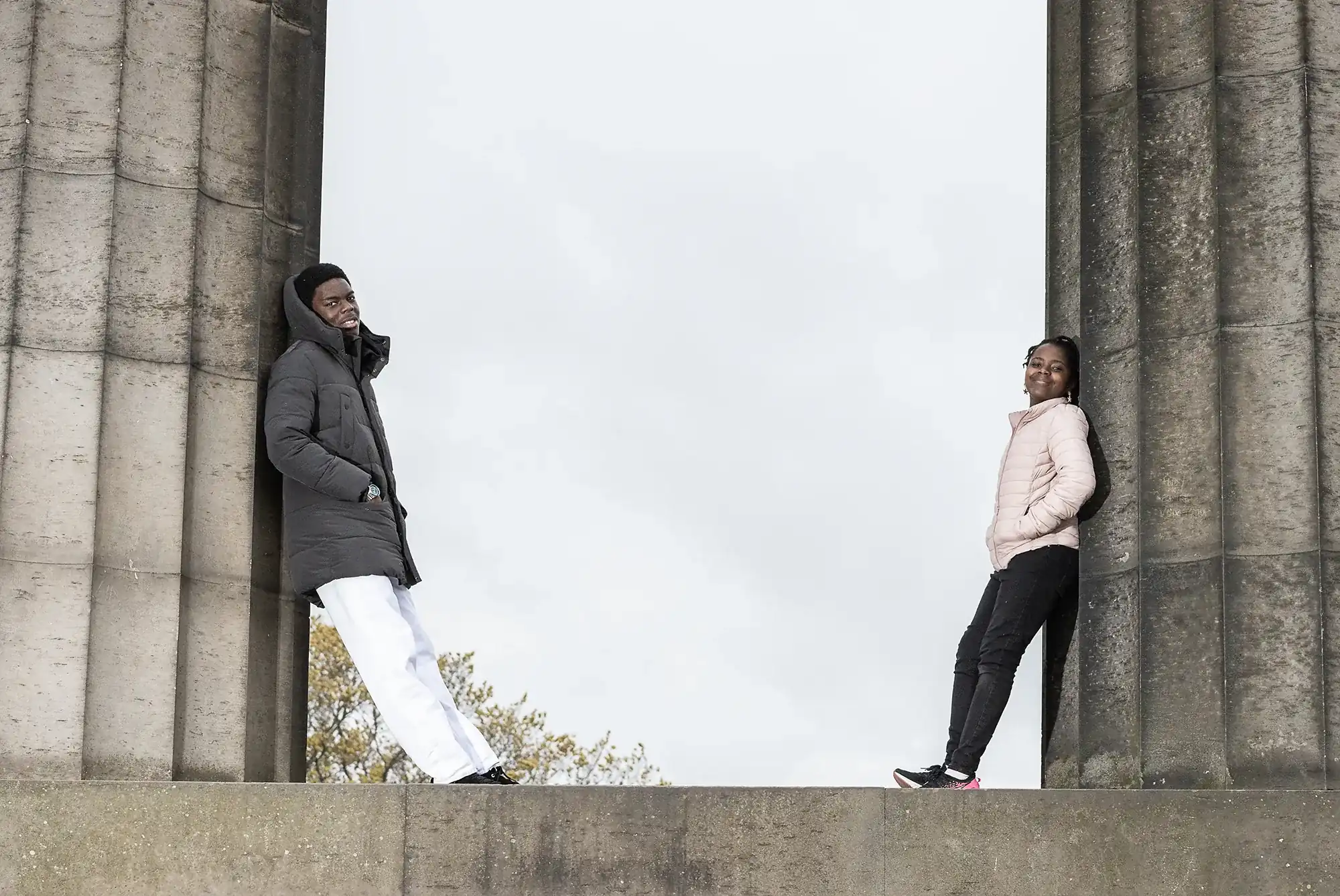 Two people stand against opposite pillars of a large stone structure. One wears a dark coat and white pants, while the other wears a light jacket and dark pants. They look towards the camera.