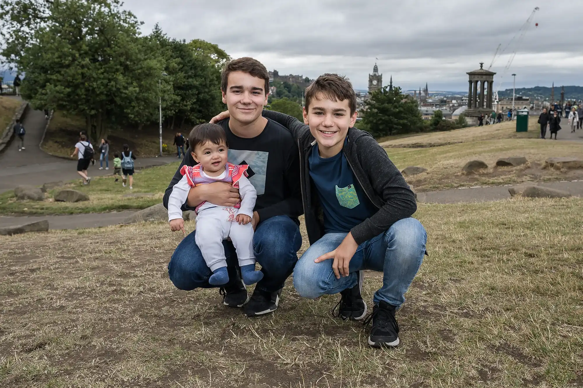 Three boys kneel on a grassy hill, with two older boys smiling and holding a baby. A park, trees, and buildings are visible in the background under a cloudy sky.