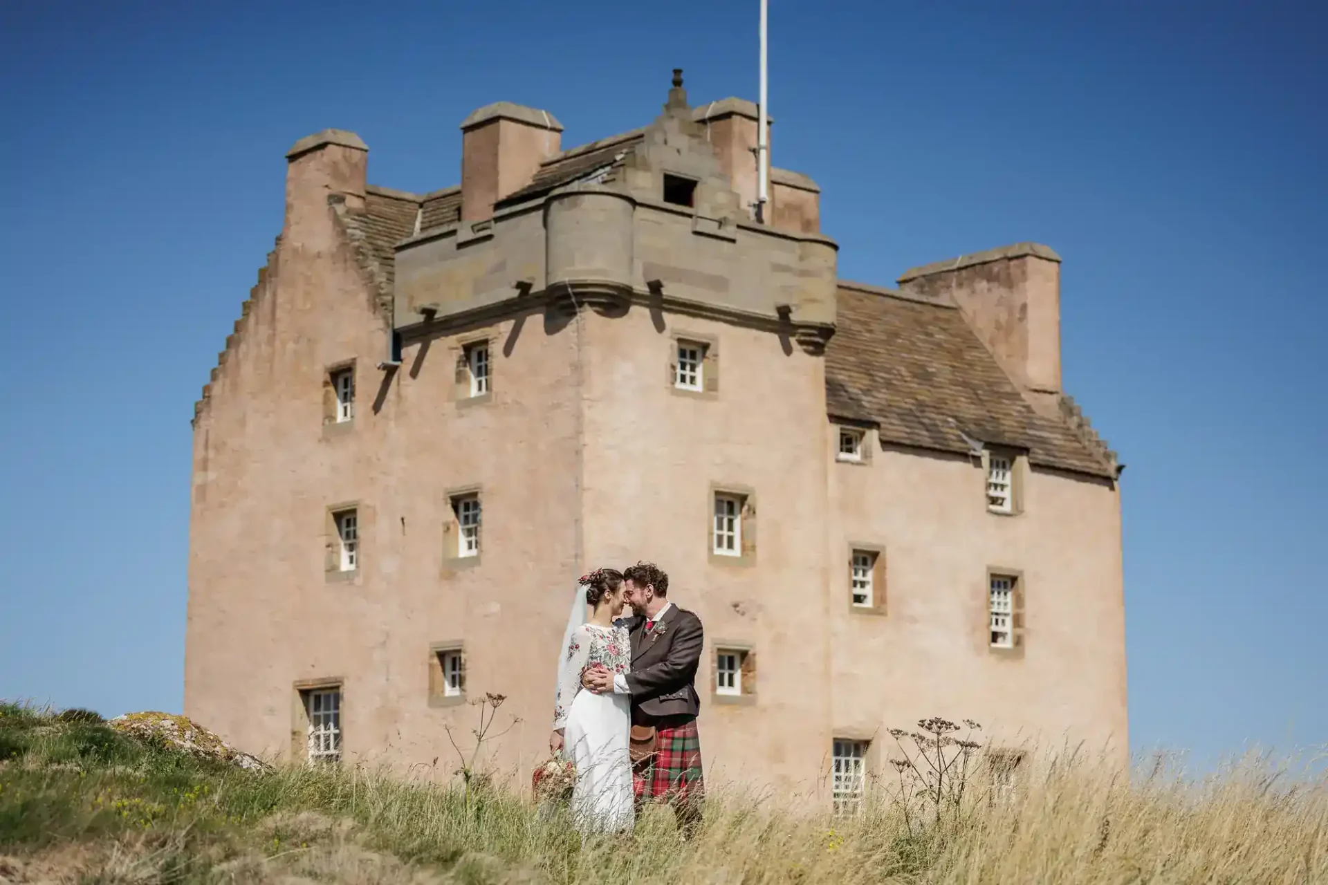 A bride and groom stand close together in front of a large historic stone building, Fenton Tower, under a clear blue sky.