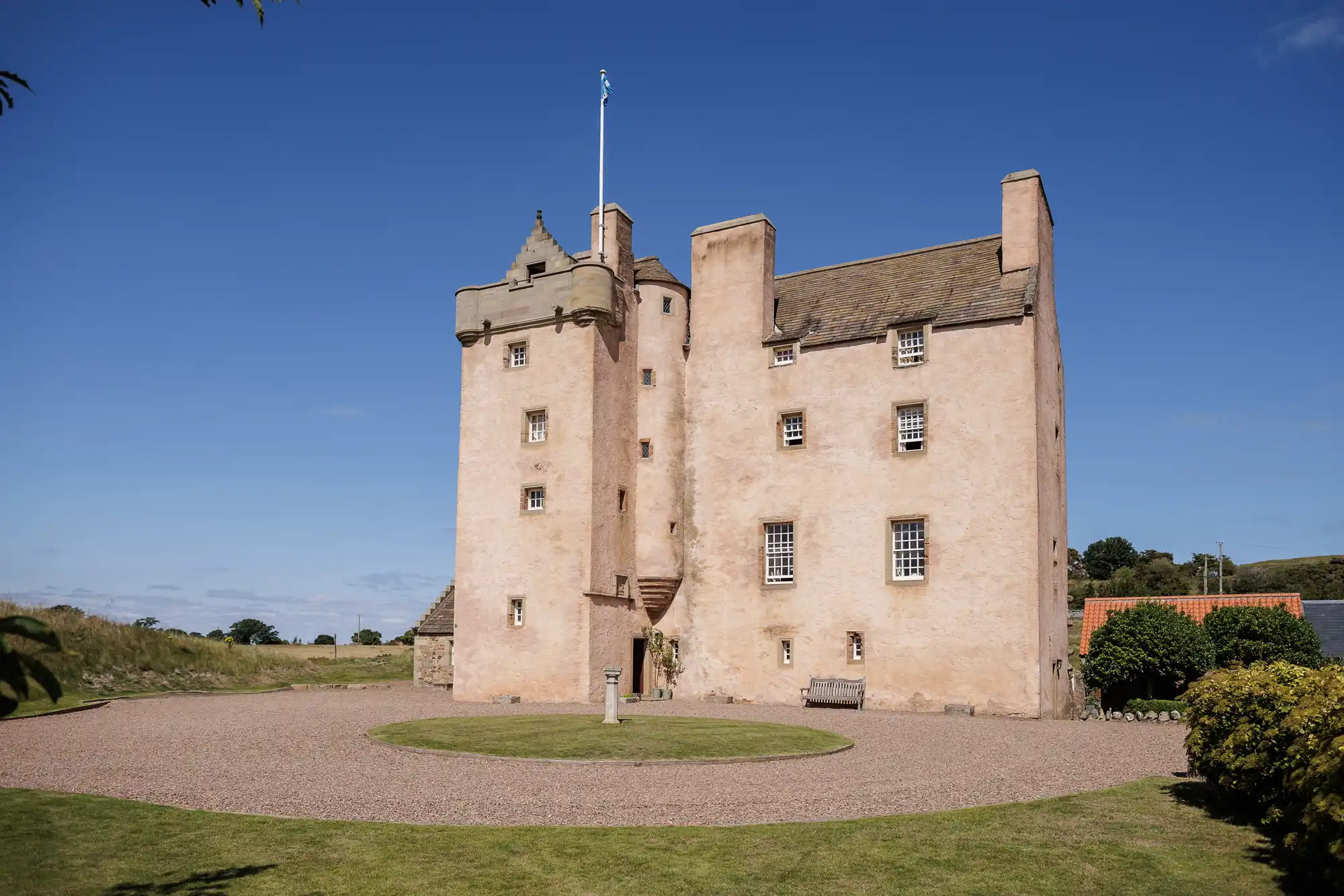 Fenton Tower is a pale stone, rectangular castle with small windows and turret sits on a gravel drive, surrounded by a circular lawn. A flag flies atop the turret. The sky is clear and blue, with green fields in the background.