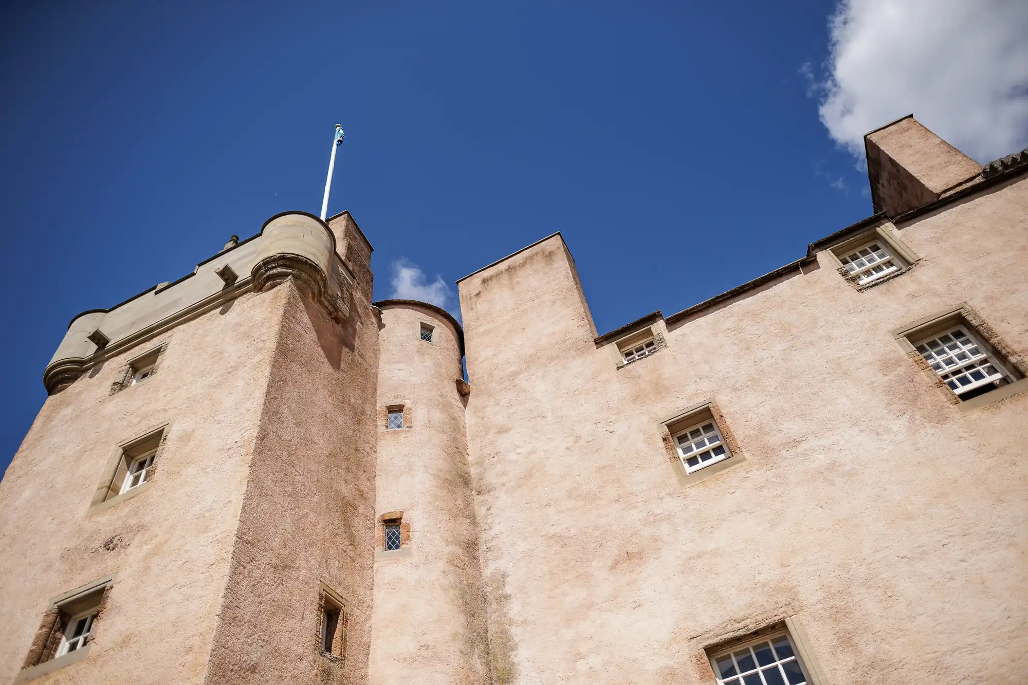 A low-angle view of a light pinkish-beige stone castle with small square windows, round turrets, and a flagpole atop one tower, set against a clear blue sky with a few clouds.