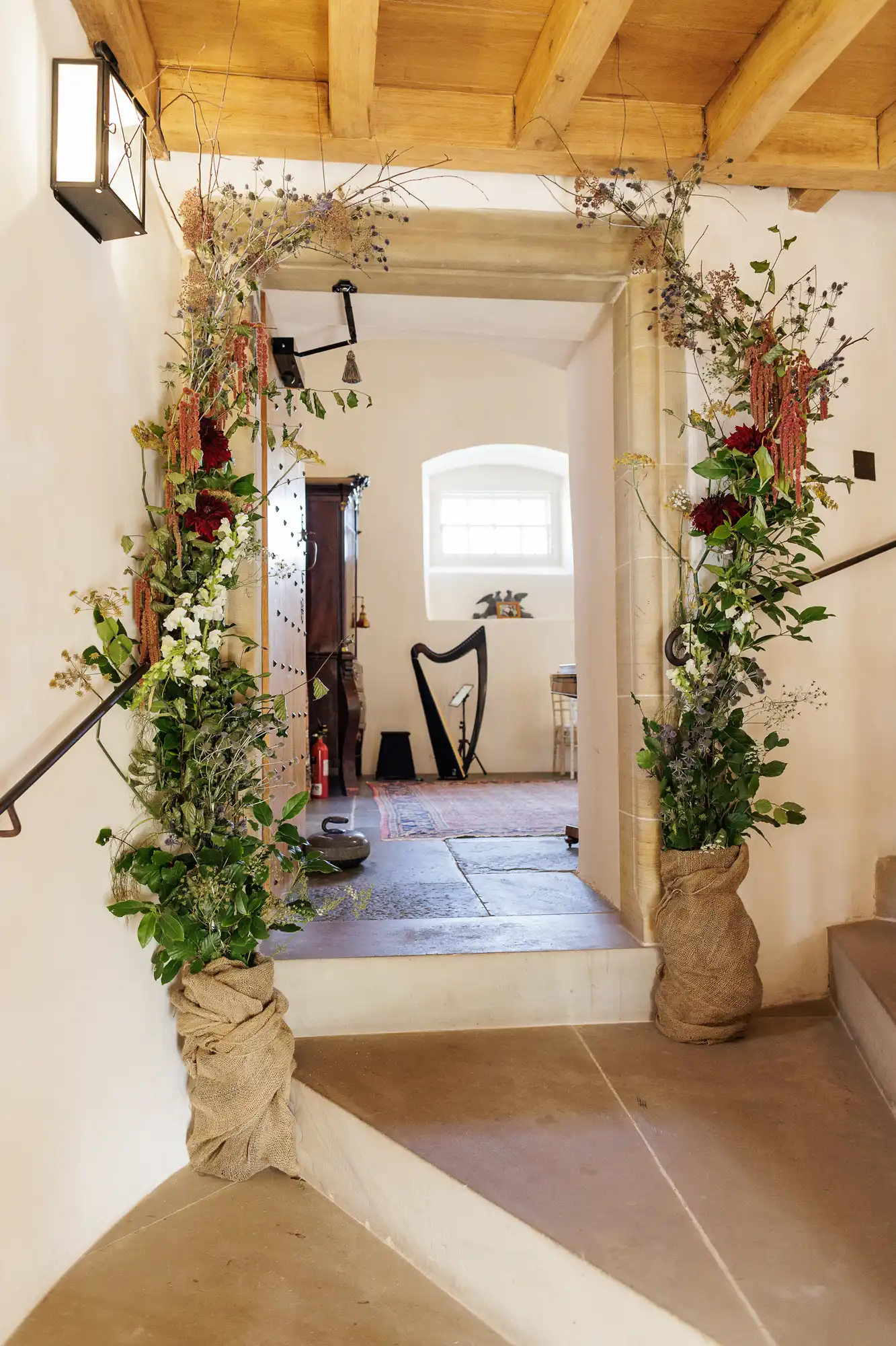 A rustic doorway decorated with tall, lush green and dried floral arrangements wrapped in hessian. Beyond the doorway, a sunlit room features a dark harp, wooden furniture, a red rug, and a large window with white walls and a stone floor.