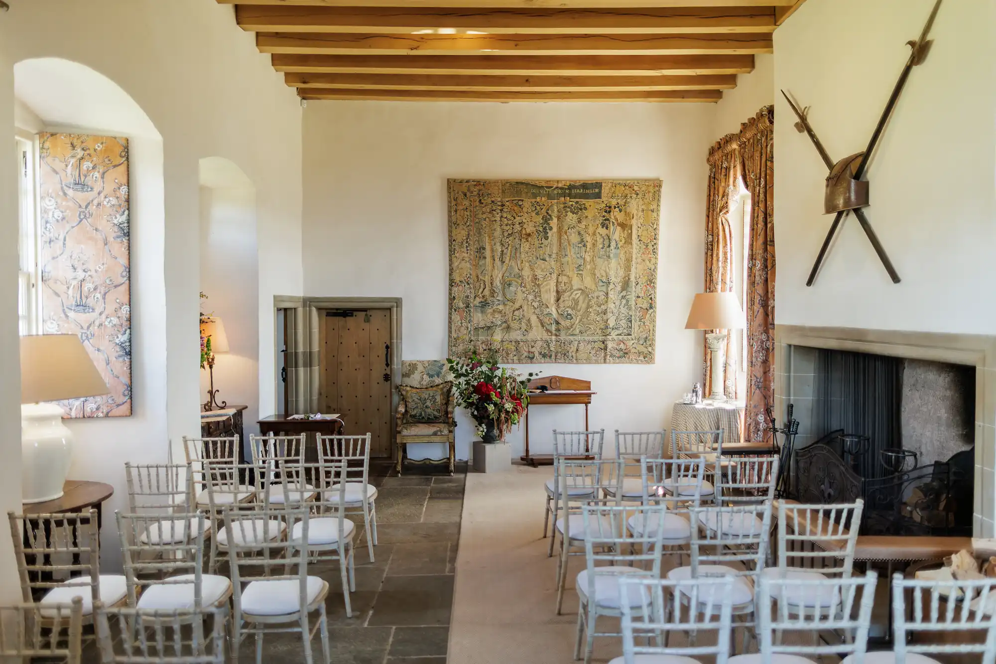 A small, elegant room set for a wedding or event with white chairs facing a lectern. The room has a stone floor, wooden beams, large fireplace, mediaeval tapestry, floral arrangements, and decorative swords crossed above the mantelpiece. Sunlight streams through windows.