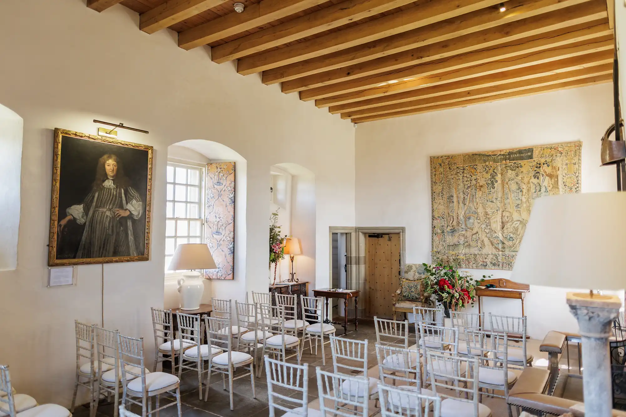 A bright room with white walls, wooden ceiling beams, and stone floor, set up for an event with rows of white chairs. Large windows, a portrait, a tapestry, lamps, and floral arrangements decorate the elegant space.