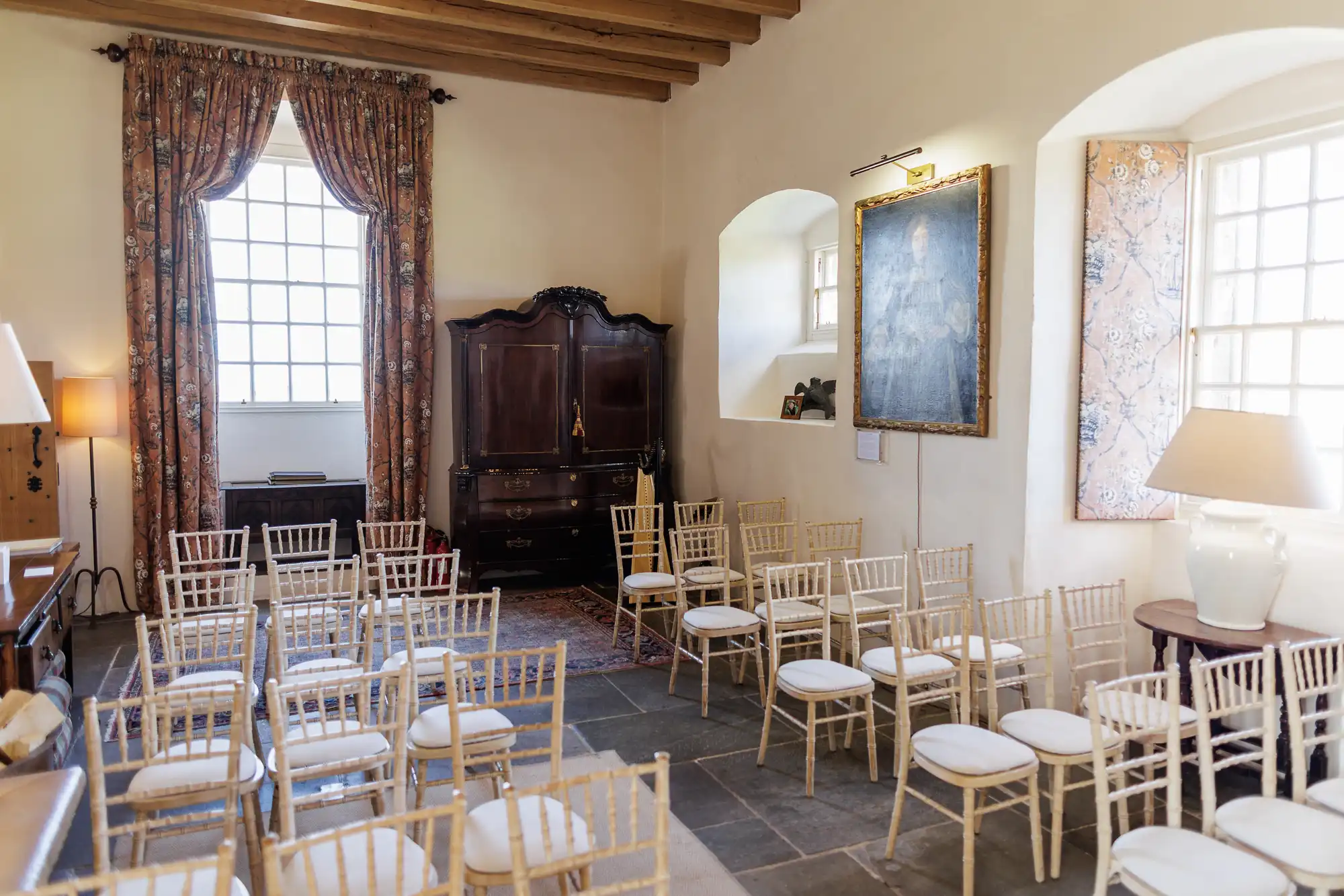 A sunlit room with stone floors and wooden beams has rows of empty white-cushioned chairs facing a wooden cupboard. Large windows with curtains, framed artwork, a piano, lamp, and rug add cosy, elegant touches to the space.