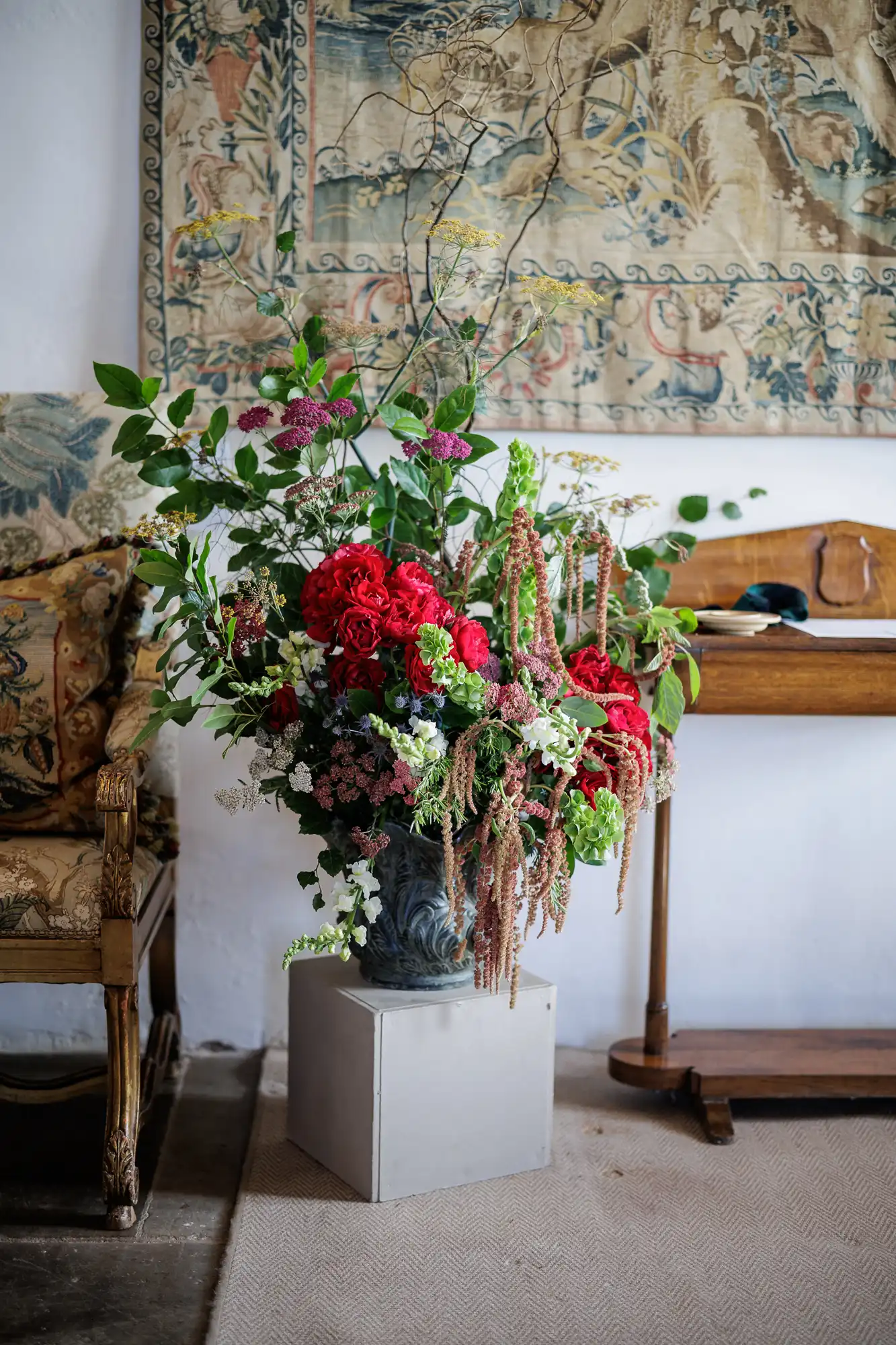 A large, ornate vase overflowing with a lush, rich floral arrangement of red roses, green hydrangeas, trailing amaranthus, and leafy branches sits atop a grey pedestal in a vintage room with tapestry, wooden furniture, and an upholstered armchair.