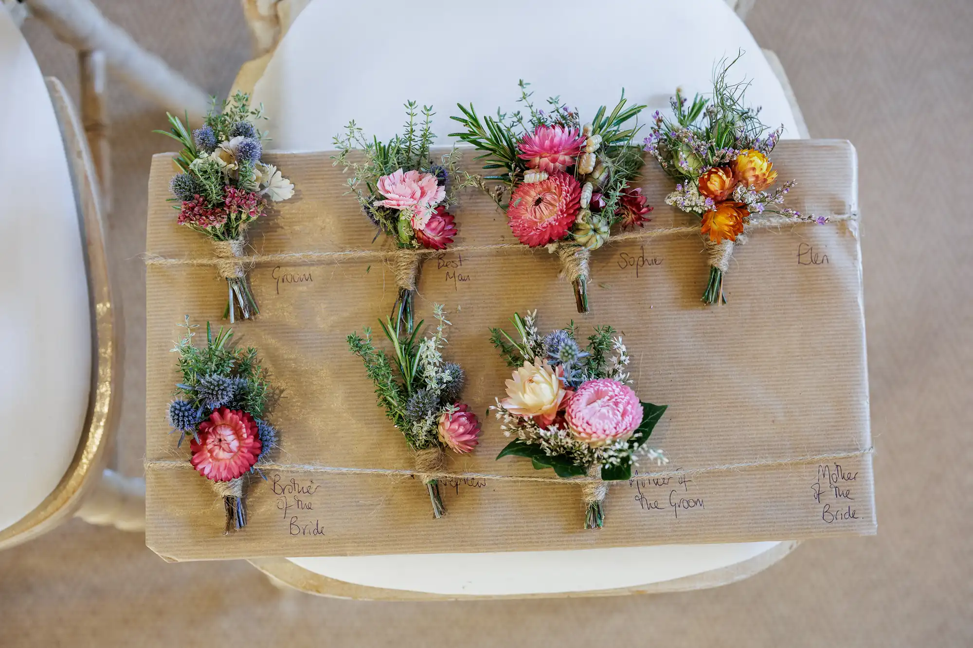 Six small, colourful floral buttonholes are arranged on brown paper, each tied with twine. Handwritten names label each buttonhole. The arrangement sits on a white cushioned chair with a second chair partially visible.