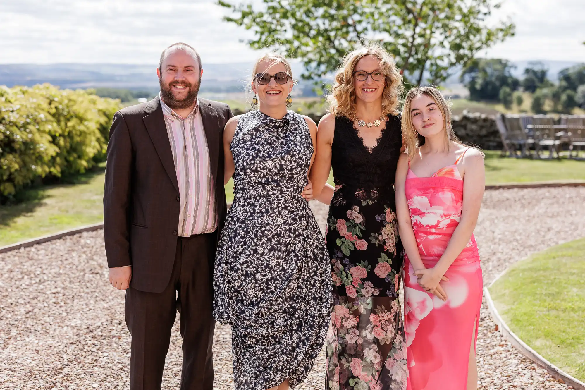 Four adults pose outdoors on a sunny day. The man wears a brown suit with a striped shirt. The three women wear floral dresses: one in black and white, one in black and pink, and one in a bright pink floral dress. Greenery and hills are in the background.