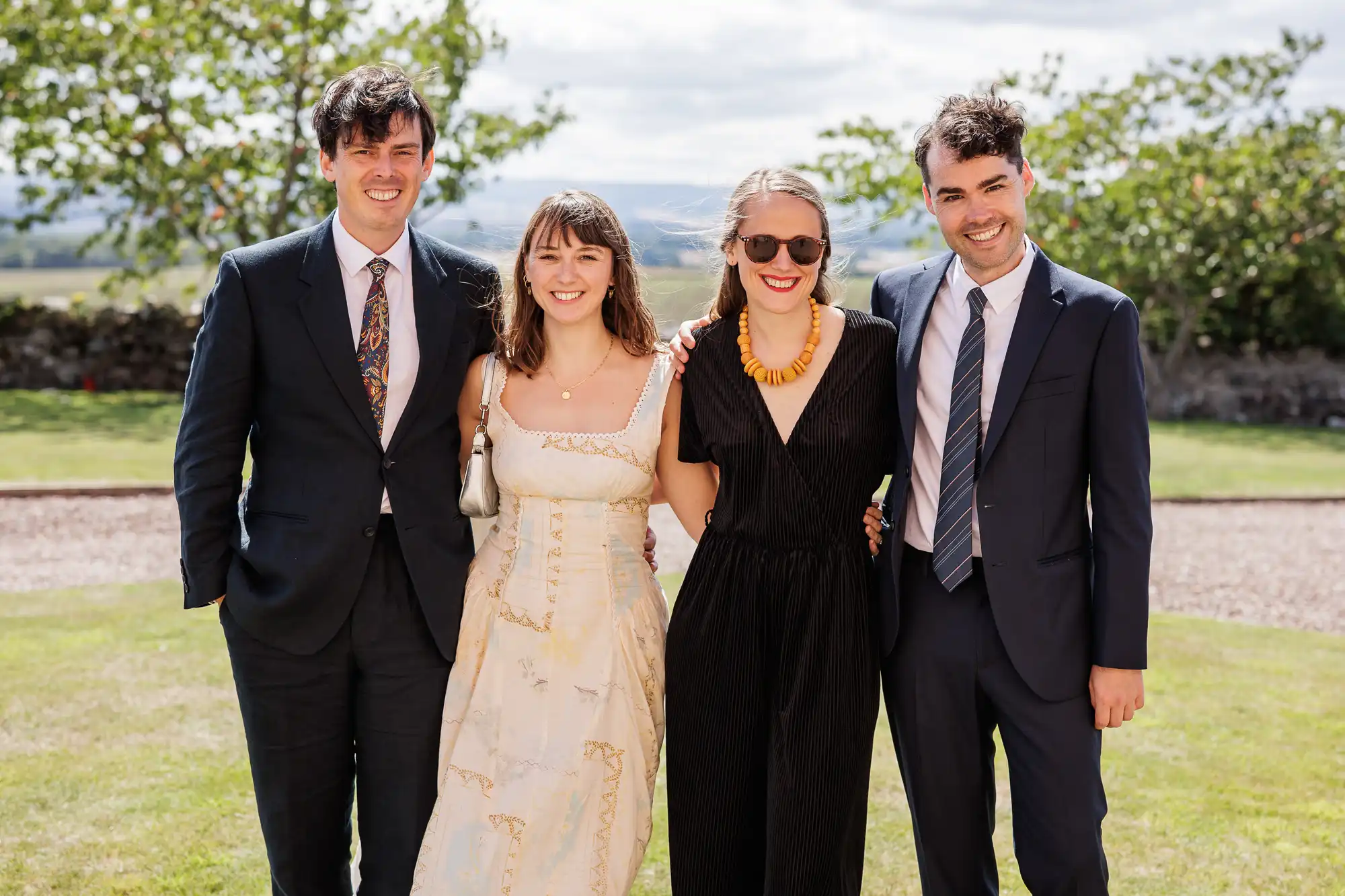 Four adults stand outside on a sunny day, smiling at the camera. Two men in suits are on either side of two women; one wears a cream dress and the other wears a black jumpsuit with sunglasses. Green grass and trees are visible in the background.