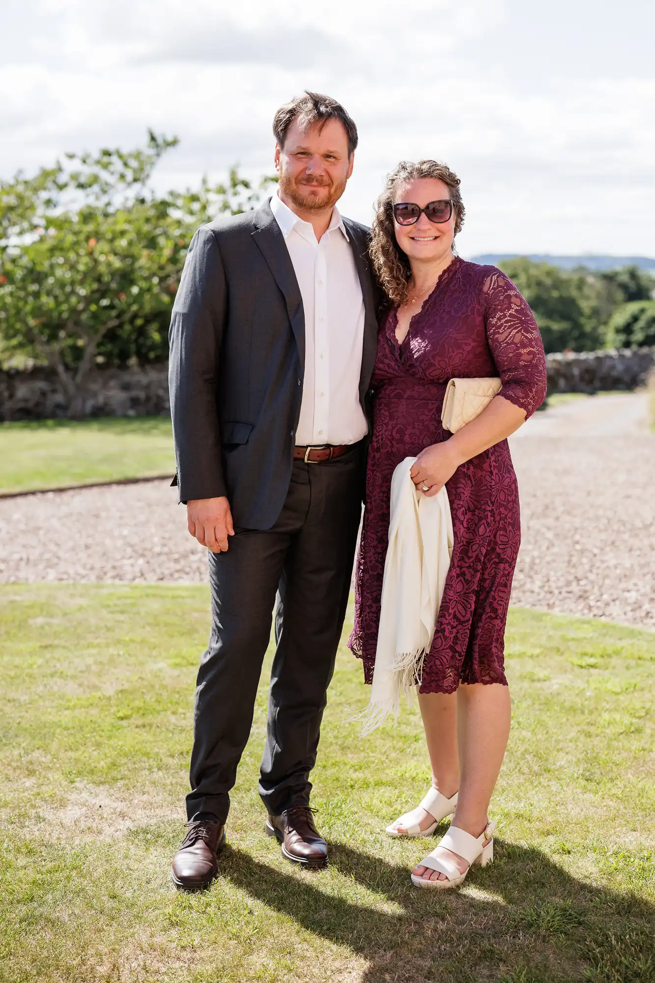 A man in a dark suit and a woman in a knee-length purple lace dress with sunglasses stand smiling together on grass, with trees and a stone path in the background. The woman holds a cream clutch and a white shawl. It is a sunny day.