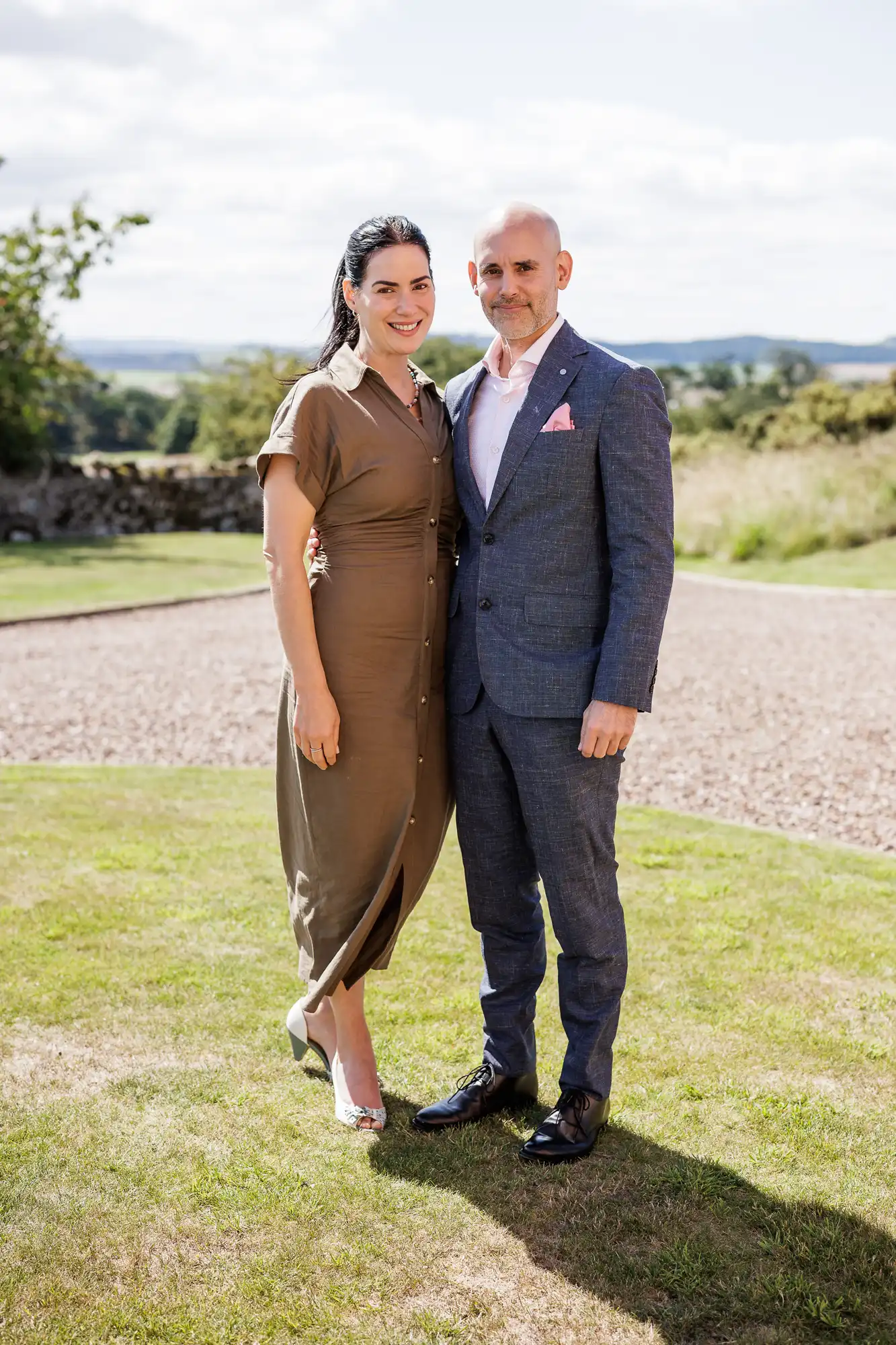 A woman in a brown buttoned dress and heels stands beside a man in a blue suit, white shirt, and black shoes. They pose together on green grass, outdoors, with trees, stone walls, and hills visible in the sunny background.