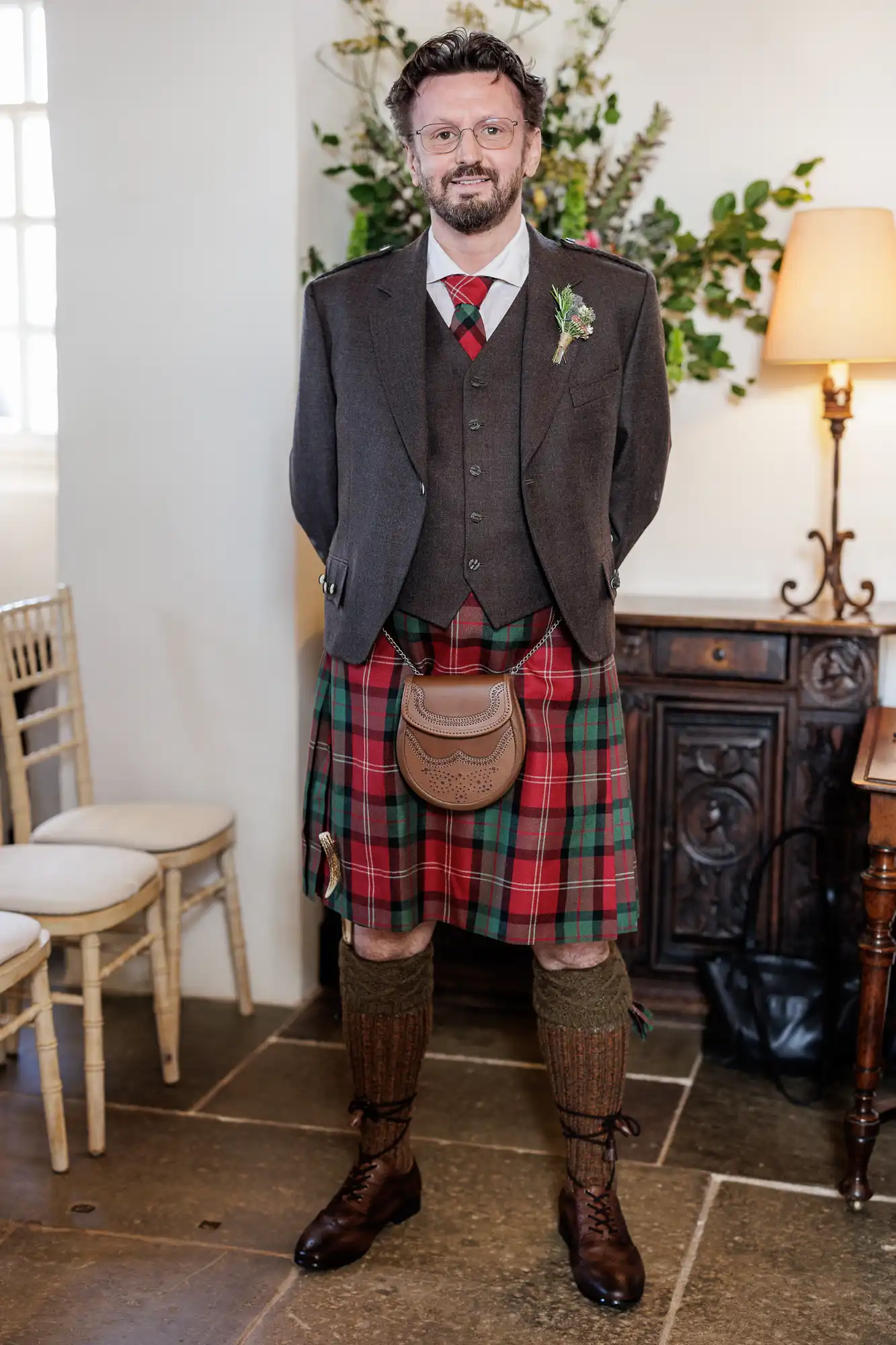A man with curly hair and glasses wears a dark jacket, waistcoat, and red tartan kilt with a brown sporran. He stands indoors on stone flooring, with green foliage and a side table, lamp, and chairs behind him, smiling at the camera.
