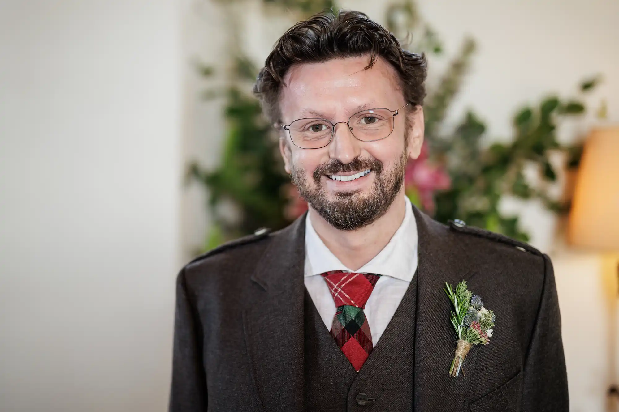 A man with glasses and a beard smiles at the camera. He wears a dark brown suit with a red and green tartan tie and a buttonhole of greenery and small flowers on his lapel. The background is softly blurred with some foliage visible.