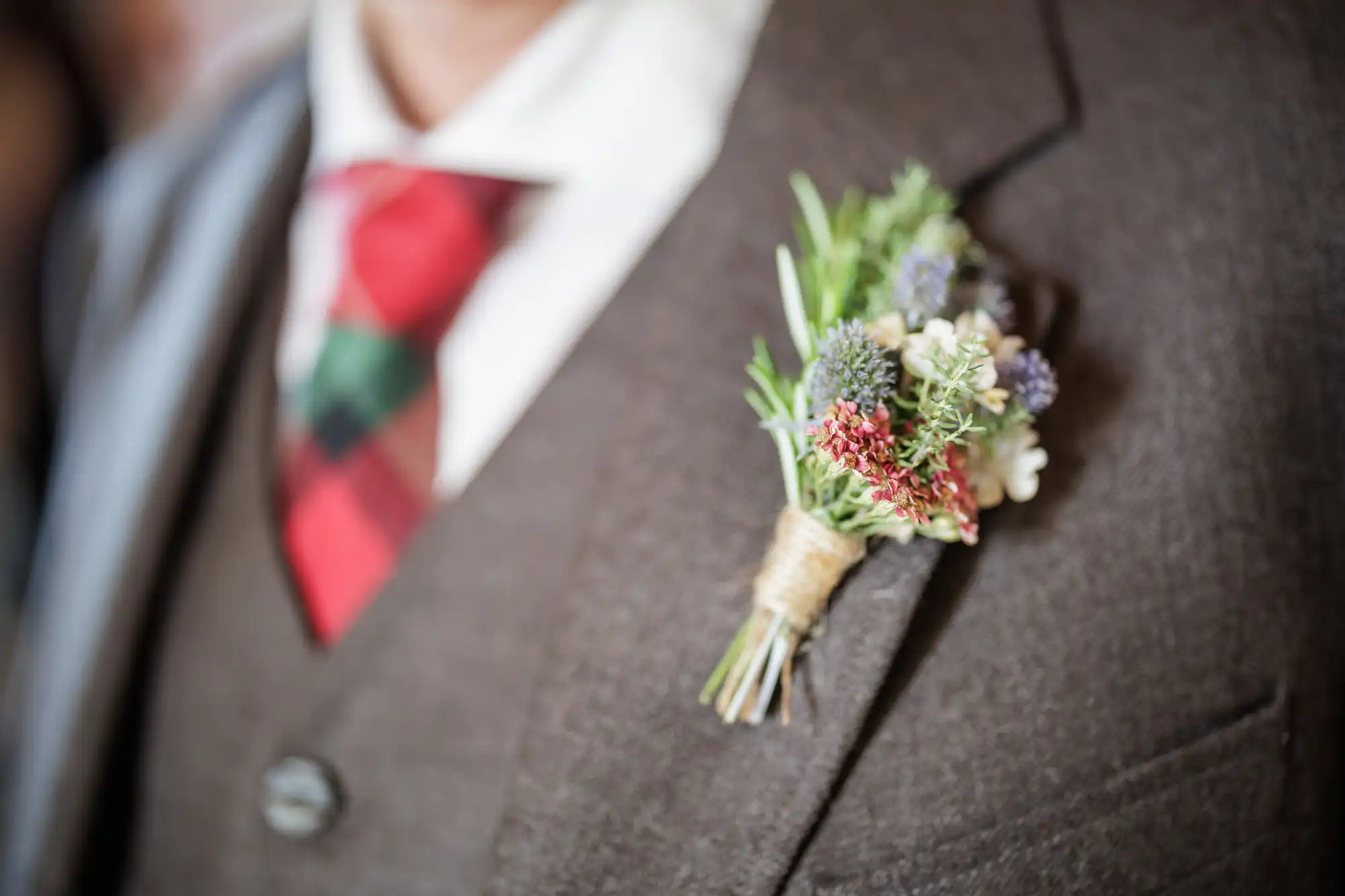 A close-up of a person wearing a brown suit, white shirt, and red-and-green checked tie. A rustic buttonhole featuring small wildflowers and greenery, tied with twine, is pinned to the lapel of the suit jacket.