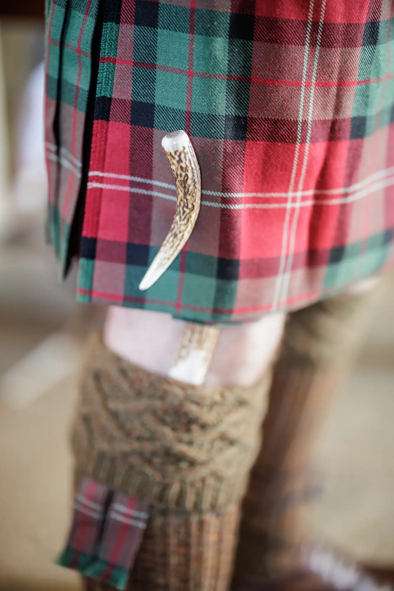 A close-up of a person wearing a red and green tartan kilt with a white-handled sgian-dubh (Scottish knife) tucked into a knitted brown sock, highlighting traditional Scottish Highland dress.