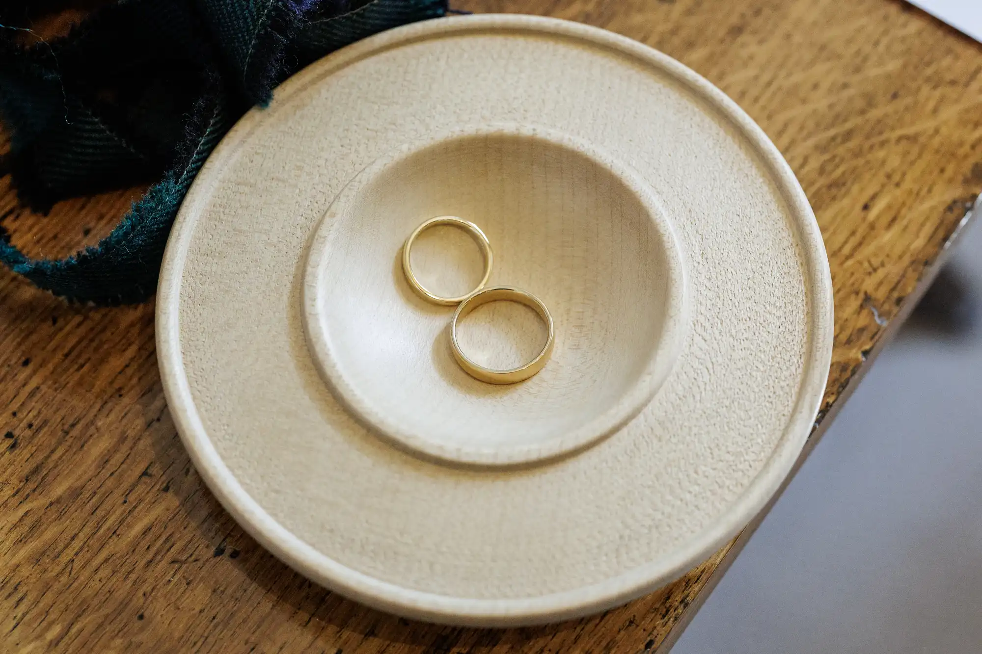 Two simple gold wedding bands rest side by side on a round, light-coloured wooden dish with a raised centre, placed on a wooden surface. A dark green fabric is partially visible in the upper left corner of the image.