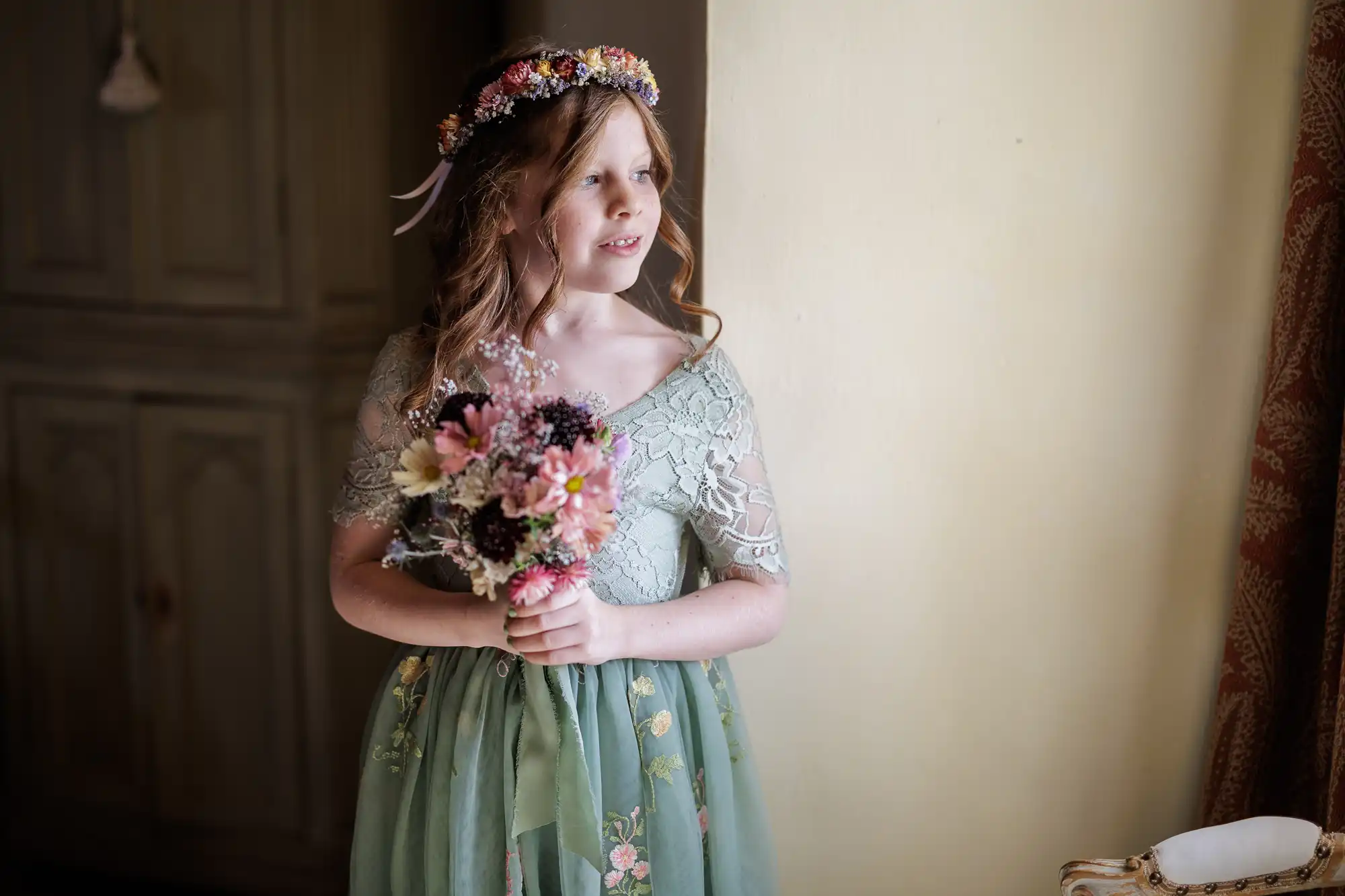 A young girl in a green, floral dress and flower crown stands indoors, holding a bouquet of pink and purple flowers. She looks towards soft sunlight coming from a window, with a gentle smile on her face. The background includes a cupboard and curtain.