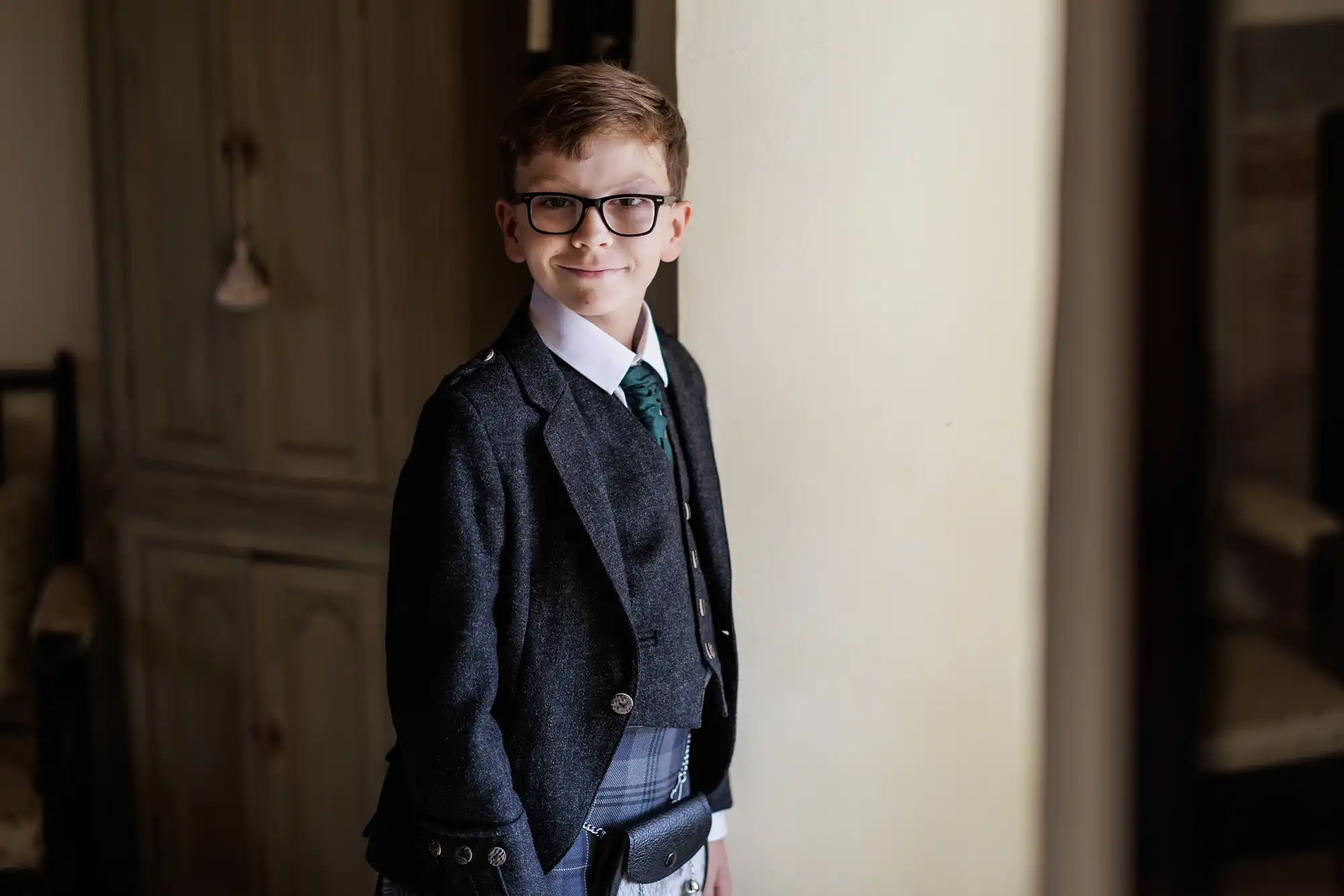 A young boy with short brown hair and glasses smiles at the camera. He wears a dark jacket, green tie, white shirt, checked kilt, and sporran, standing indoors next to a cream-coloured wall and wooden cupboard.