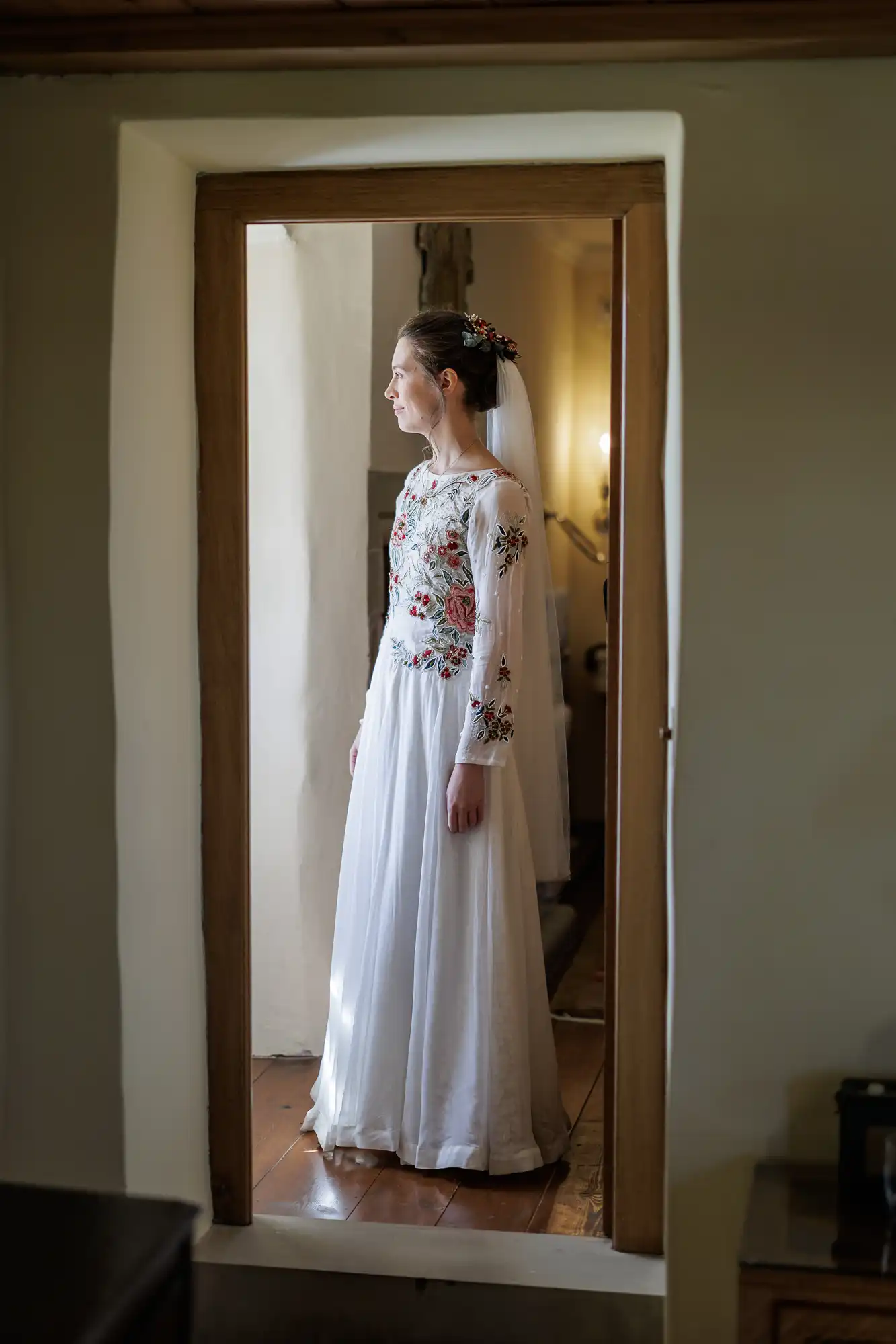 A bride in a long white dress with floral embroidery stands in a doorway, gazing to the left. She wears a veil with small flowers in her hair. Soft indoor light illuminates her profile, creating a serene and elegant atmosphere.