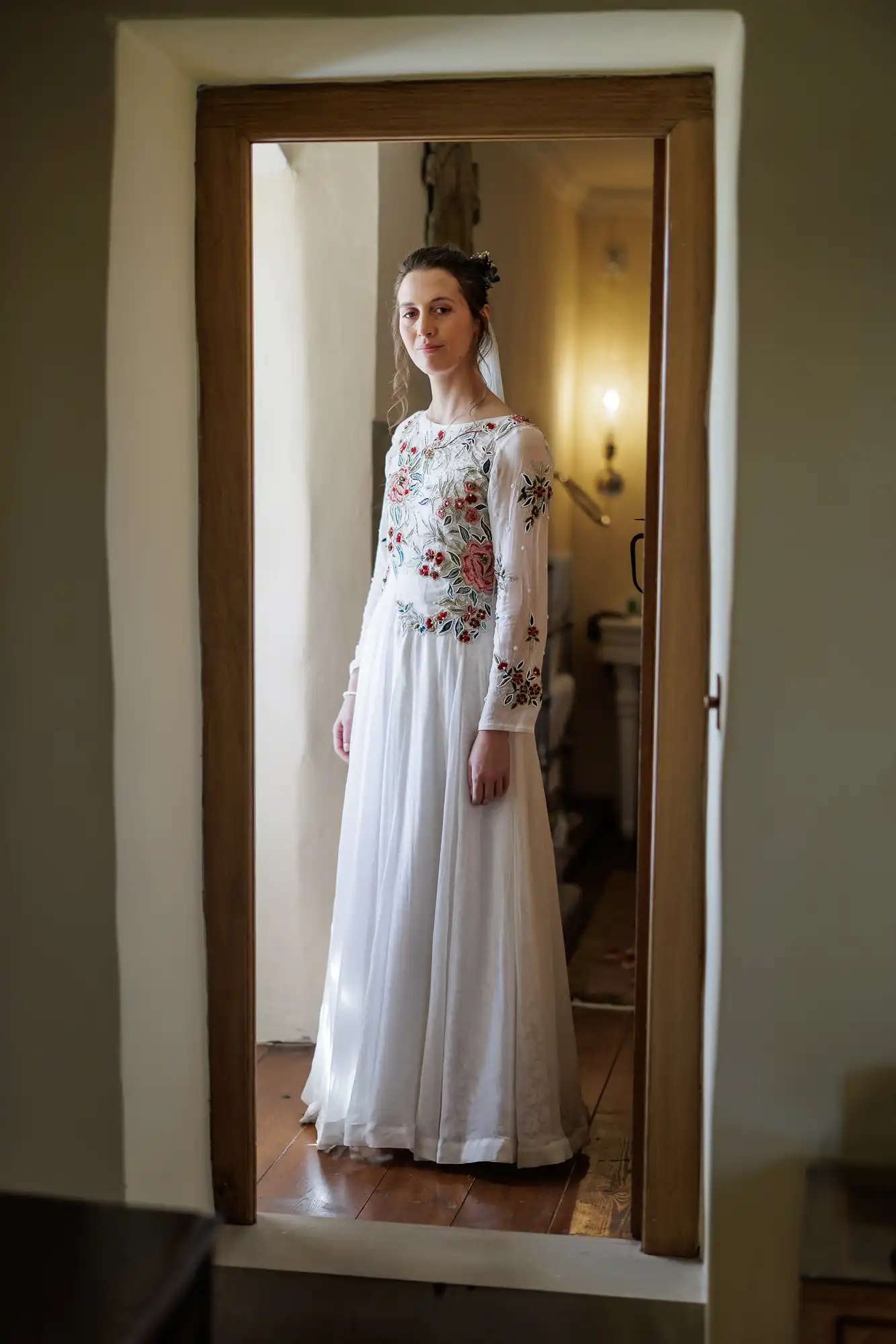 A woman in a long, flowing white dress with colourful floral embroidery on the bodice and sleeves stands framed in a doorway, looking gently at the camera. The room behind her is softly lit, with light-coloured walls and wooden floors.