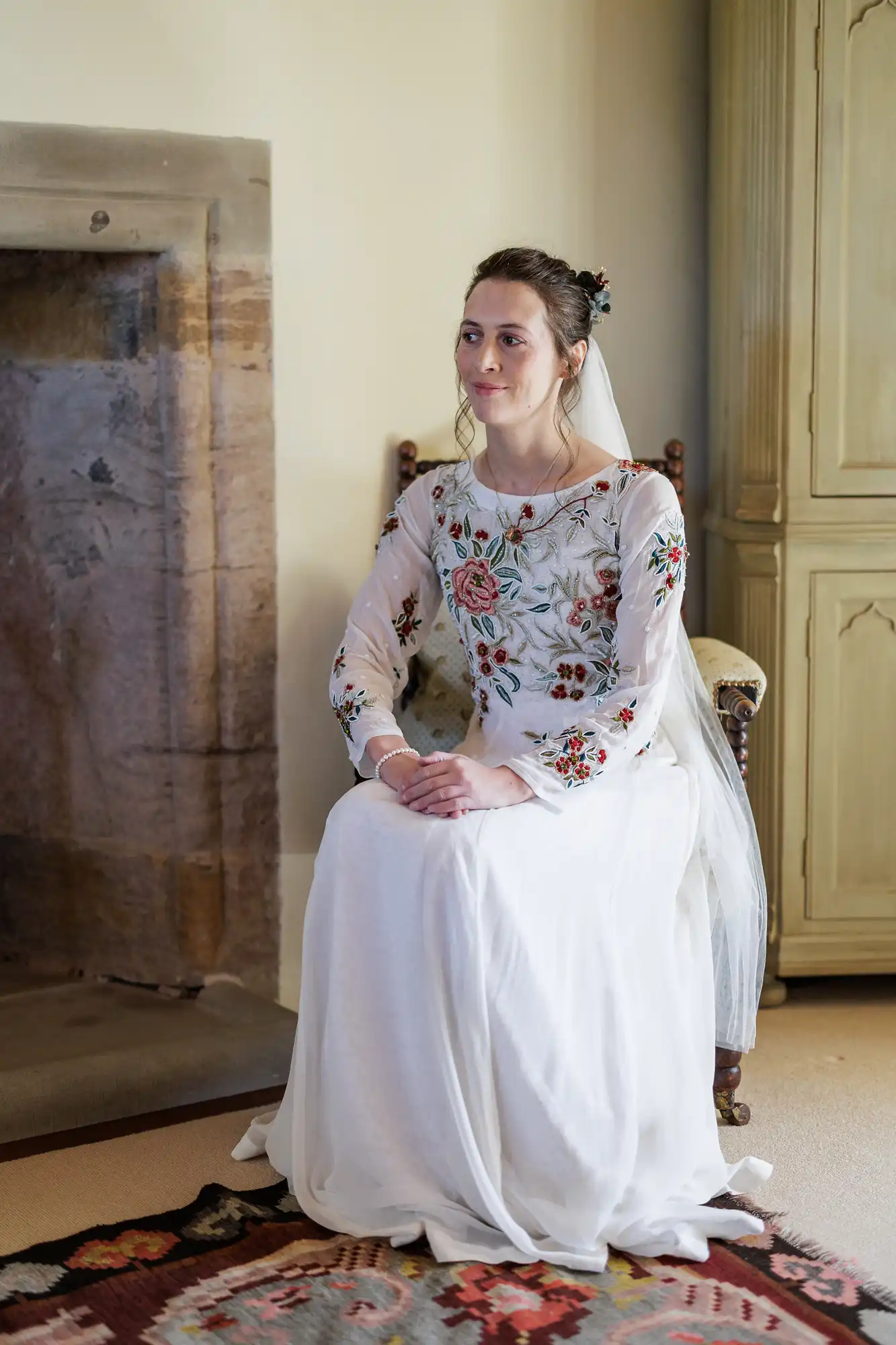 A woman in a white wedding dress with colourful floral embroidery sits on a wooden chair by a stone fireplace. She has a veil, her hair is up, and she looks slightly to the side with a calm expression. An ornate rug is on the floor.