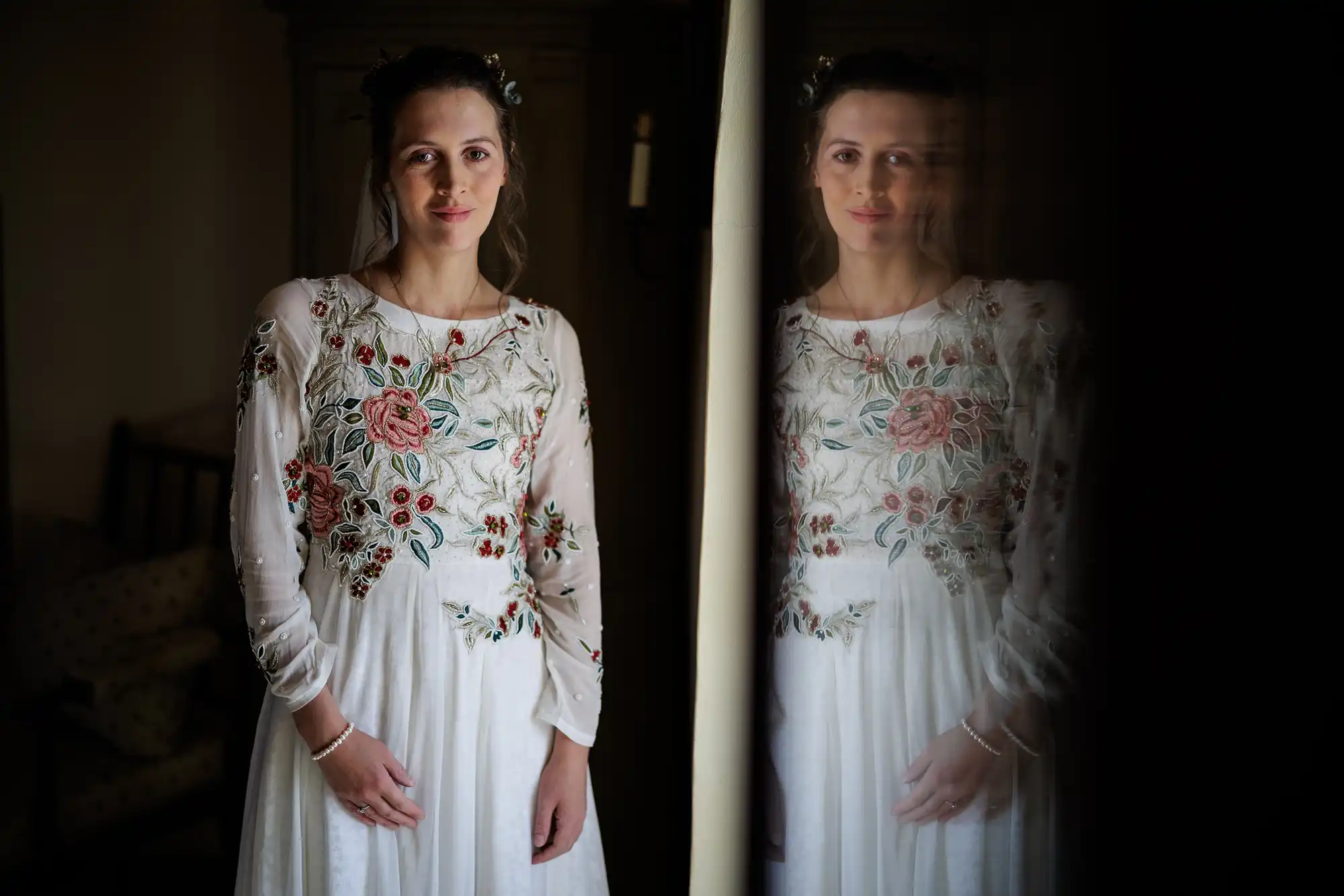 A woman in a white dress with colourful floral embroidery stands indoors next to a reflective surface, creating a soft, slightly blurred reflection of her. She has light skin, brown hair, and a gentle expression, with delicate jewellery on her wrist and neck.