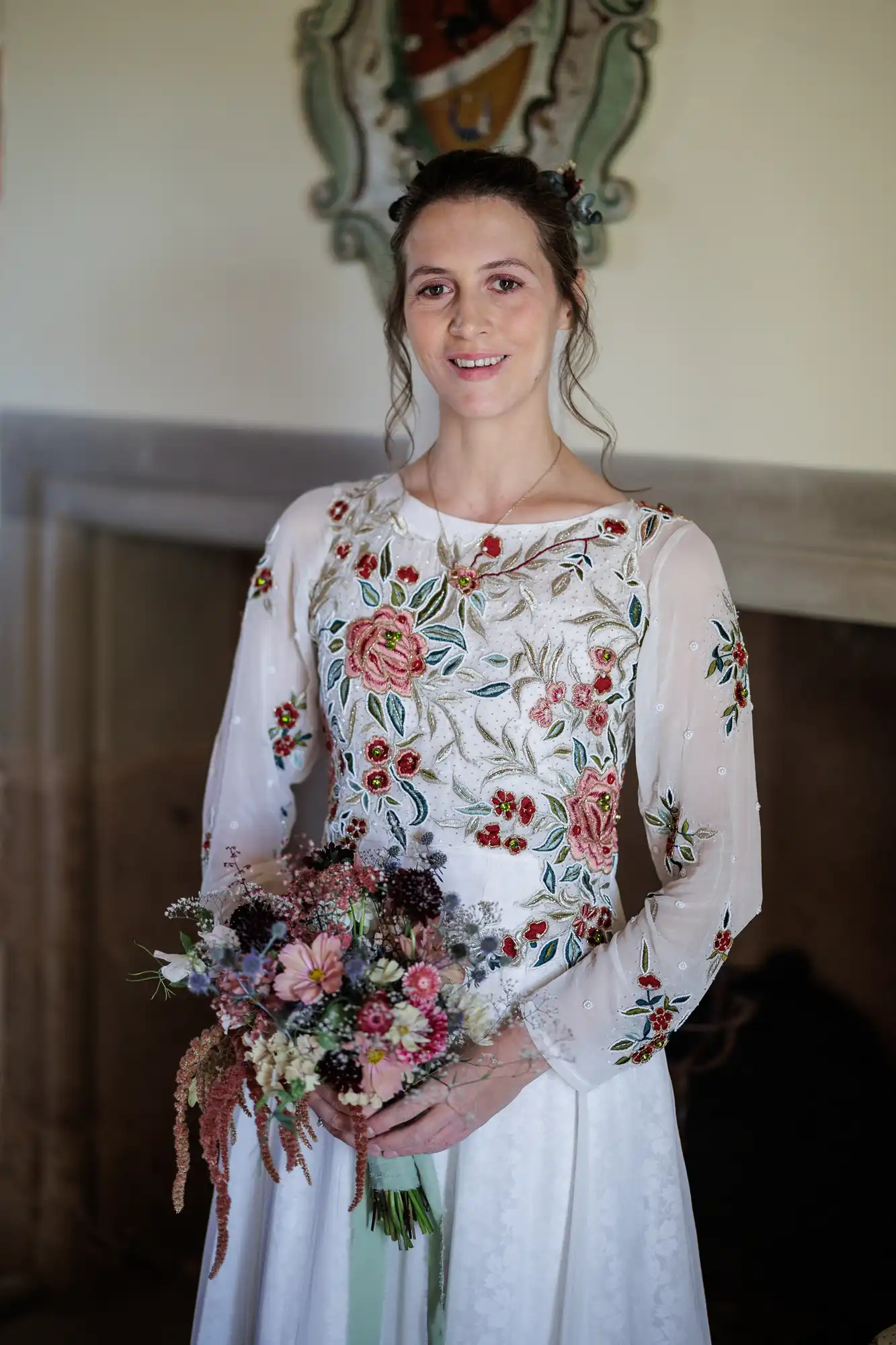 A woman in a white dress with floral embroidery holds a bouquet of assorted flowers. She has light skin, brown hair pulled back, and smiles softly. The background is indoors, with a fireplace and a decorative wall ornament visible behind her.