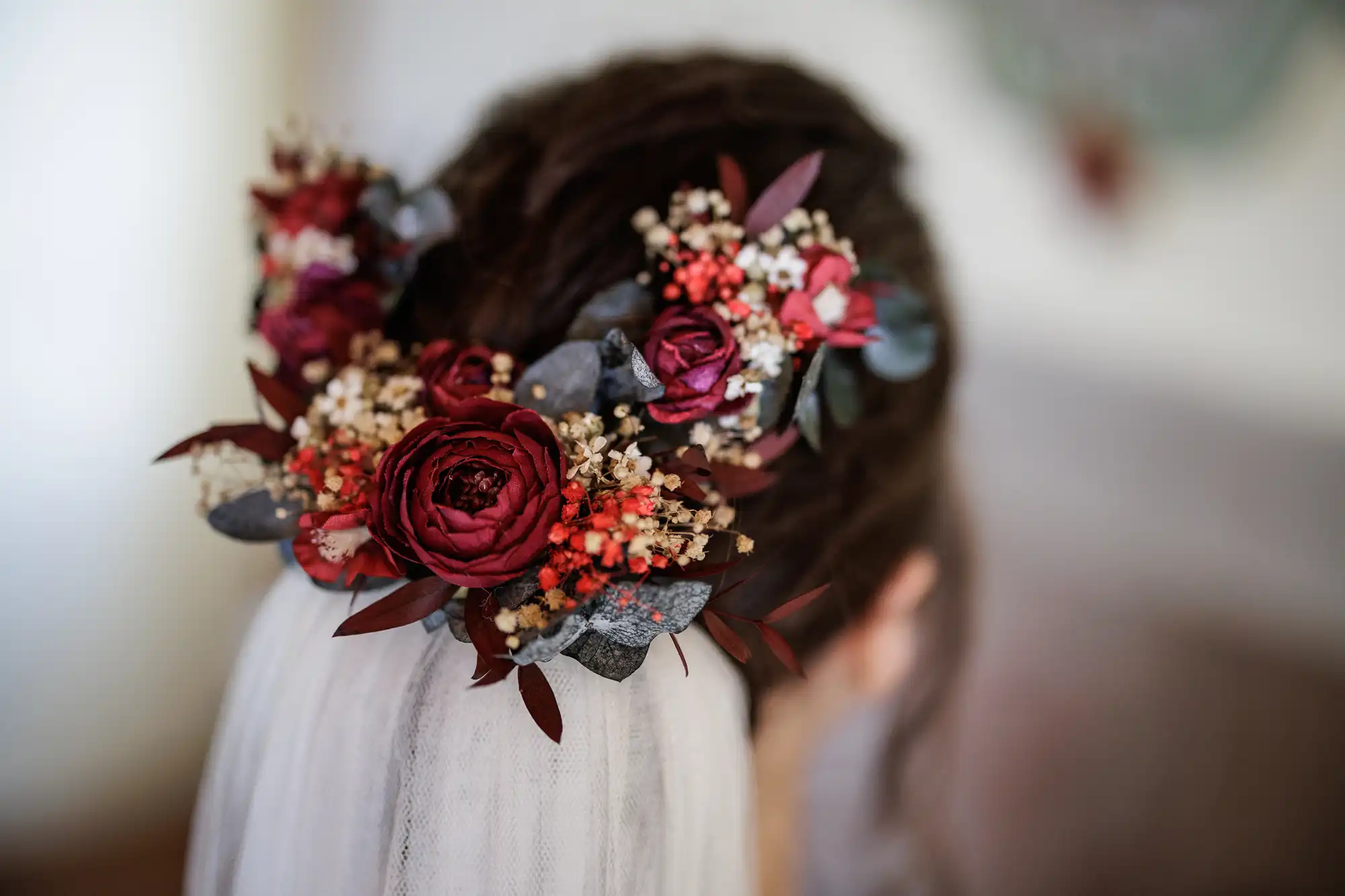 A close-up of a woman's hair adorned with a bridal hairpiece featuring deep red and burgundy flowers, baby's breath, and dark green leaves. A white veil is attached underneath the floral arrangement, and the background is softly blurred.