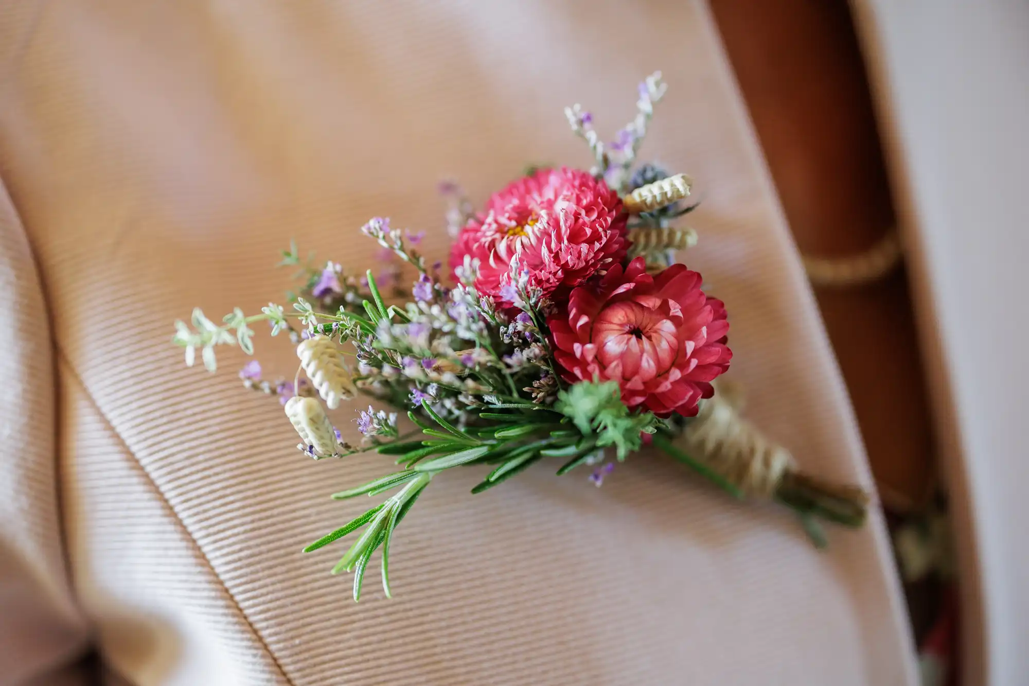 A close-up of a pink and lavender buttonhole with small flowers and green leaves pinned to a pale, textured jacket, worn over a brown top. The buttonhole is neatly wrapped at the stem with twine.